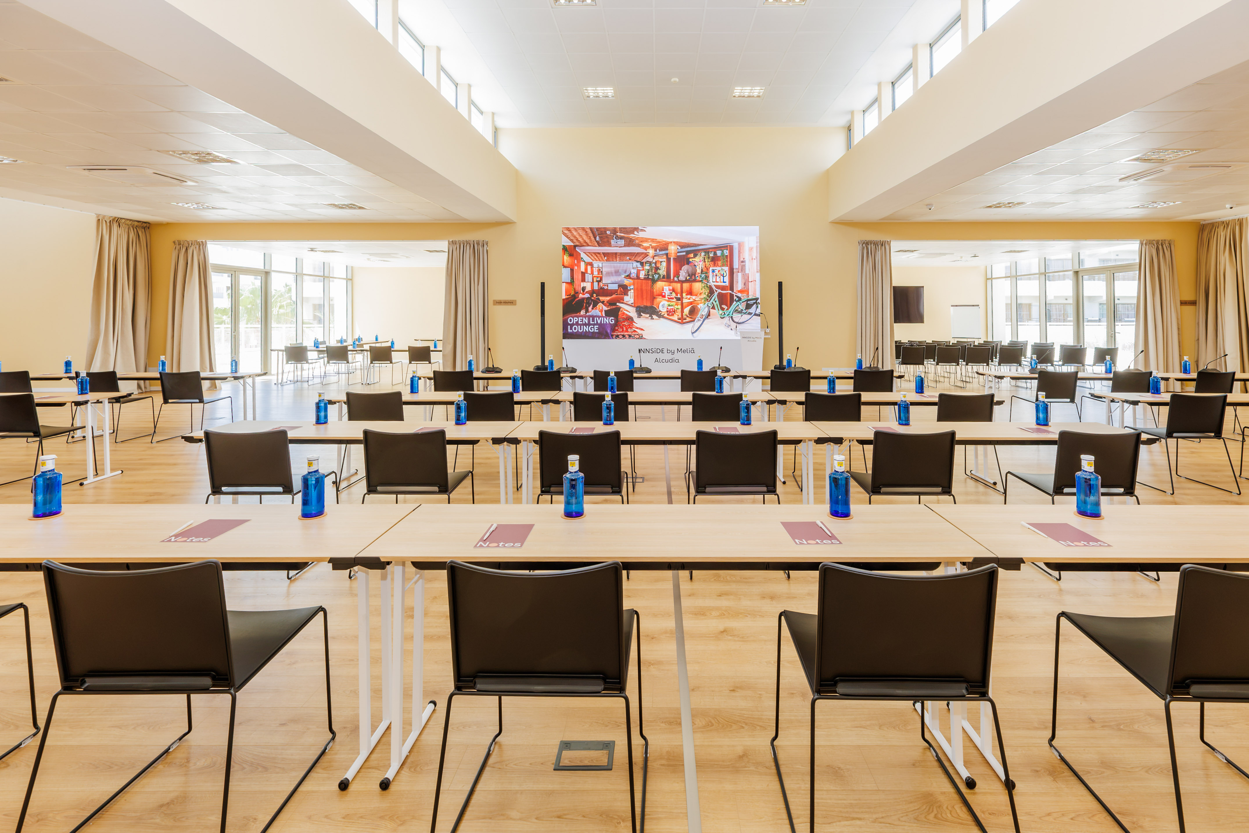 Hotel conference room with tables, chairs, water, and large brand screen.