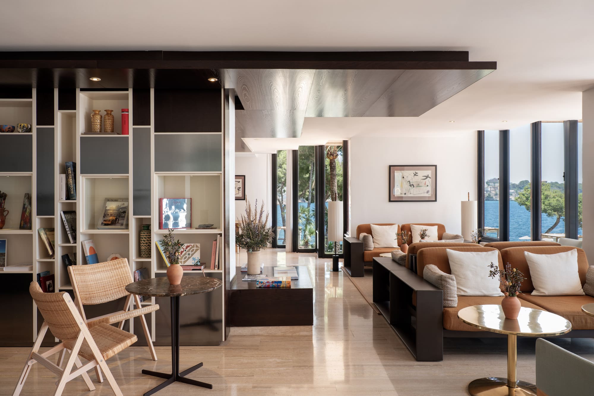 Modern hotel lounge with sea view, stylish tan sofas, and decorative bookshelves.