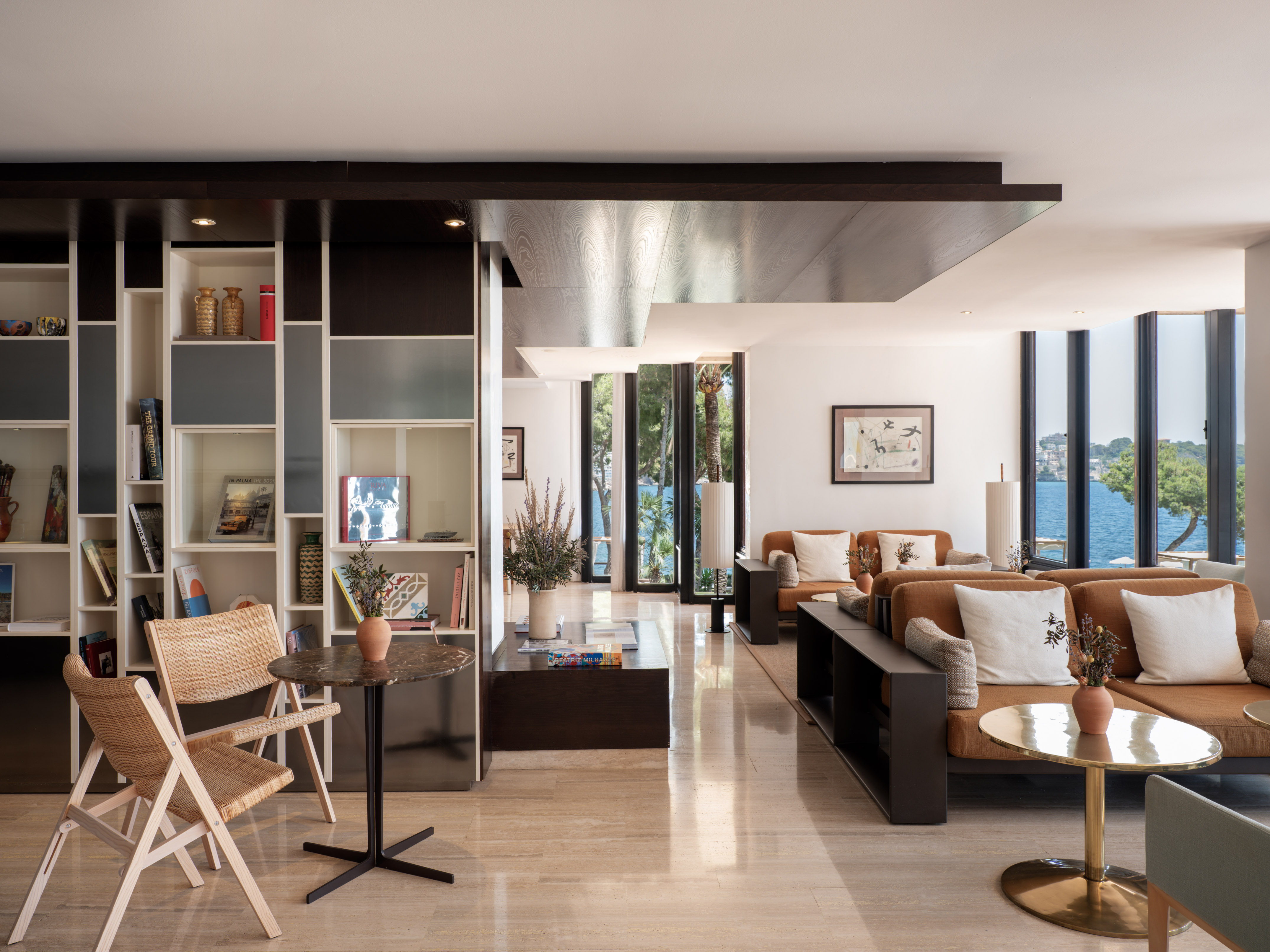 Modern hotel lounge with sea view, stylish tan sofas, and decorative bookshelves.