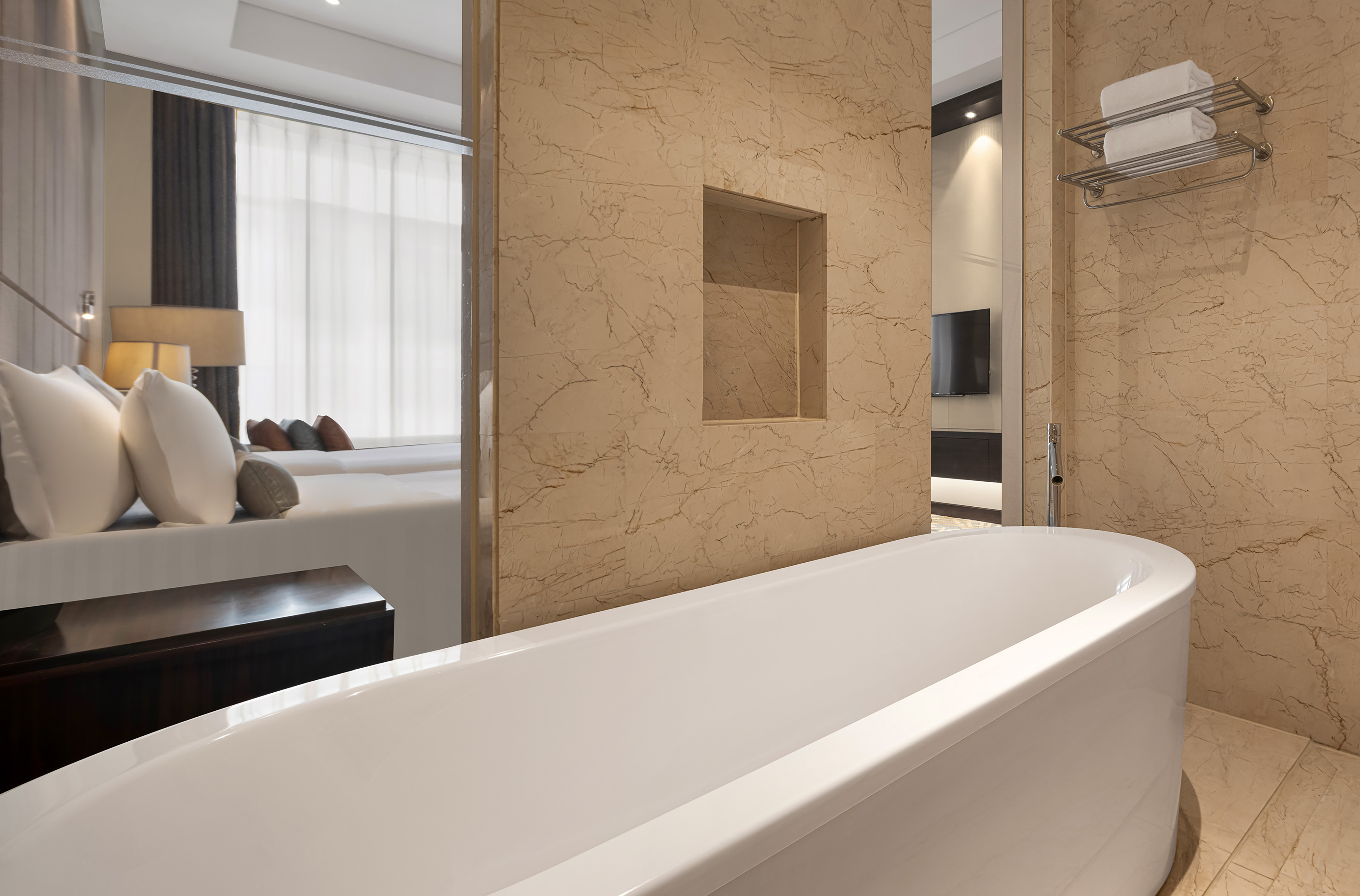 Modern hotel bathroom with white bathtub, marble walls, and bedroom view.