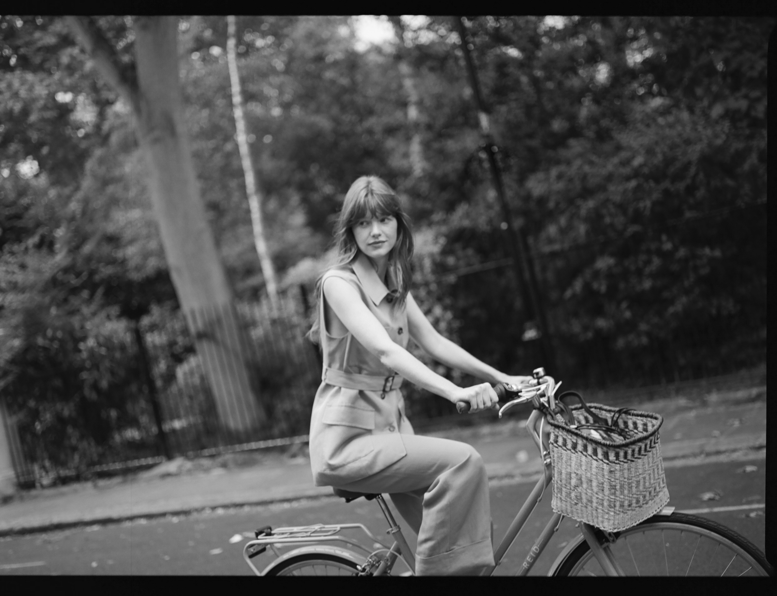 Black and white: Woman in jumpsuit rides bicycle with front basket on a street.
