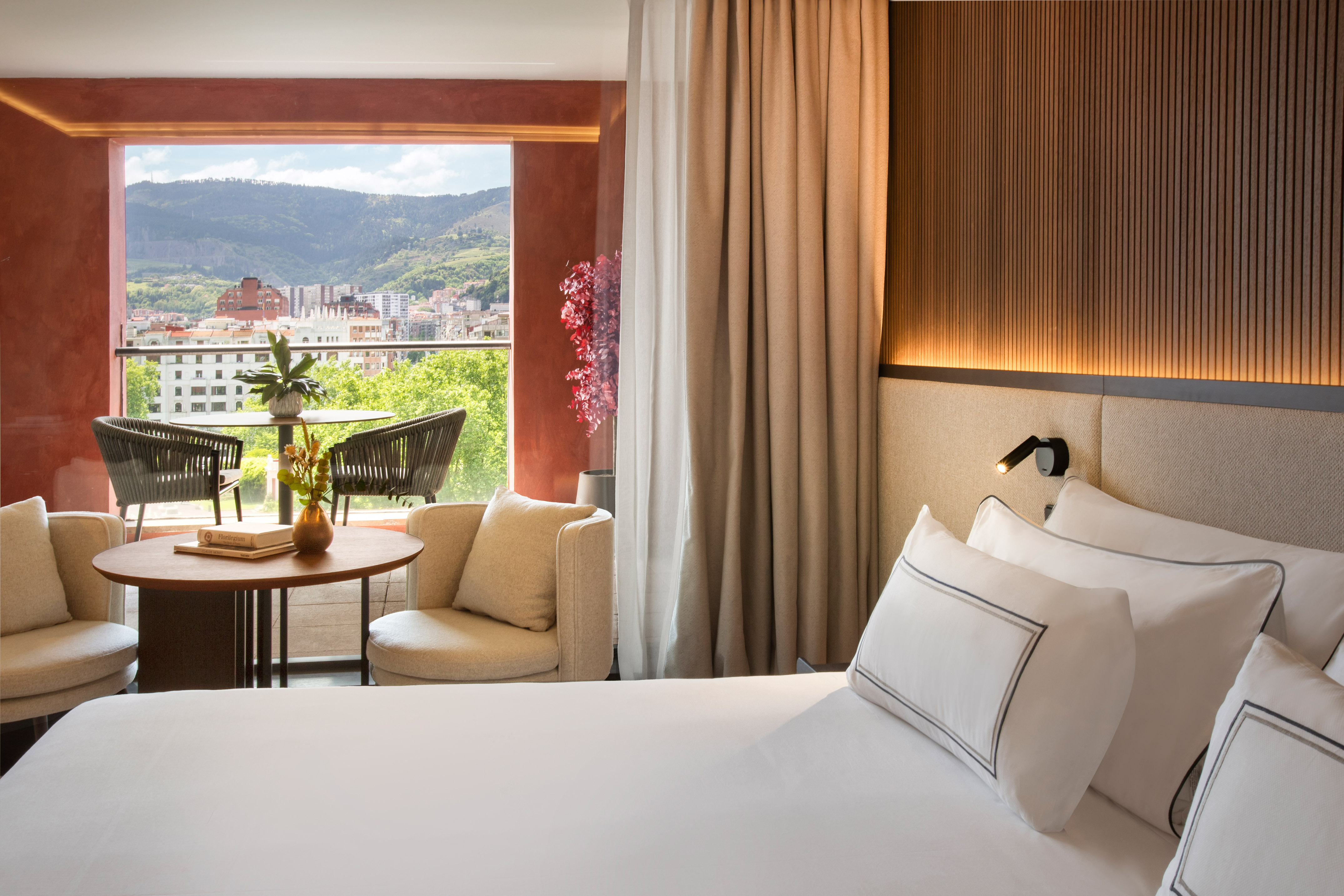 Modern hotel room with balcony, outdoor furniture, and mountain city view.