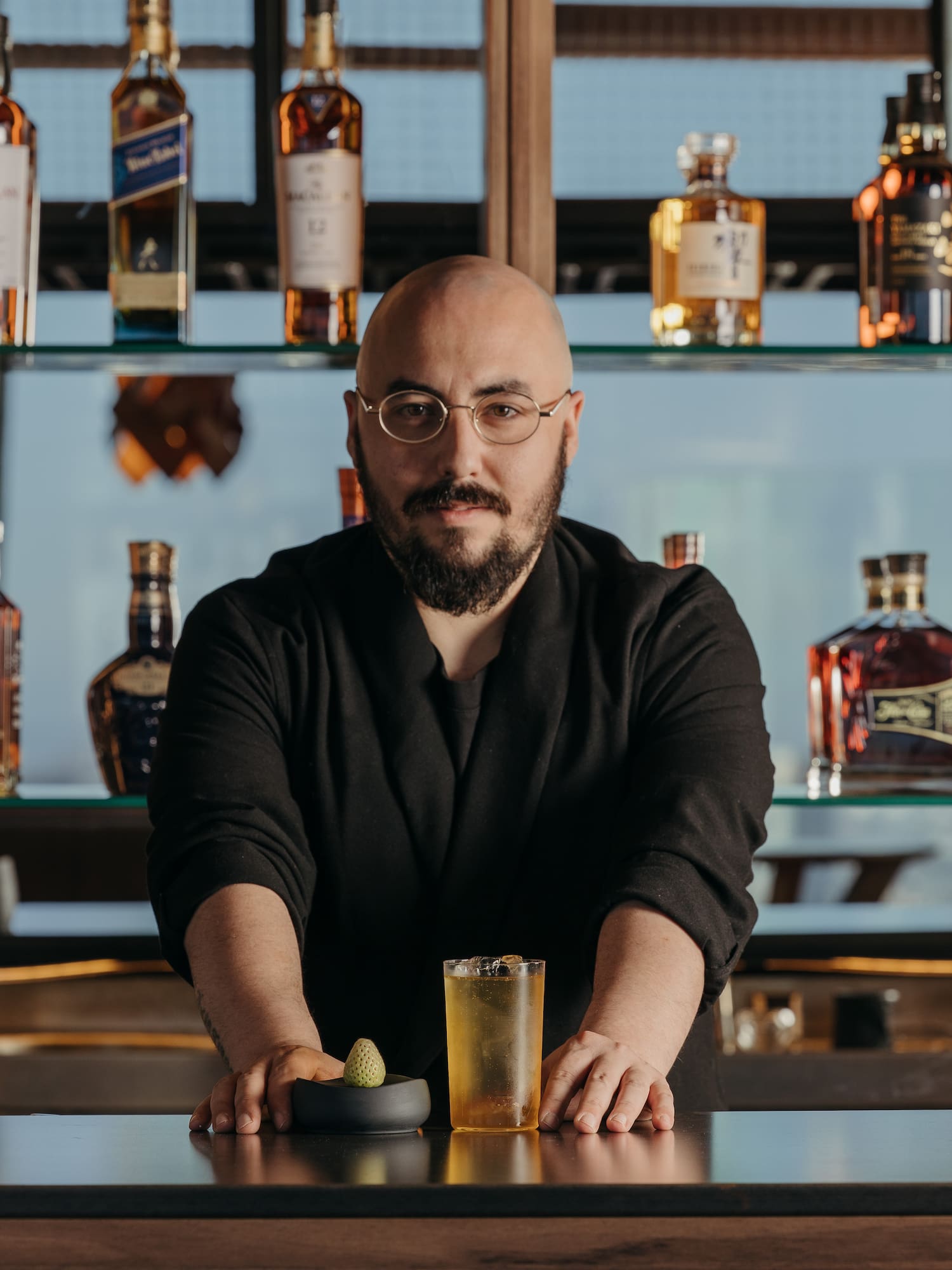 Barman barbudo con gafas presenta cóctel en barra con botellas de whisky.