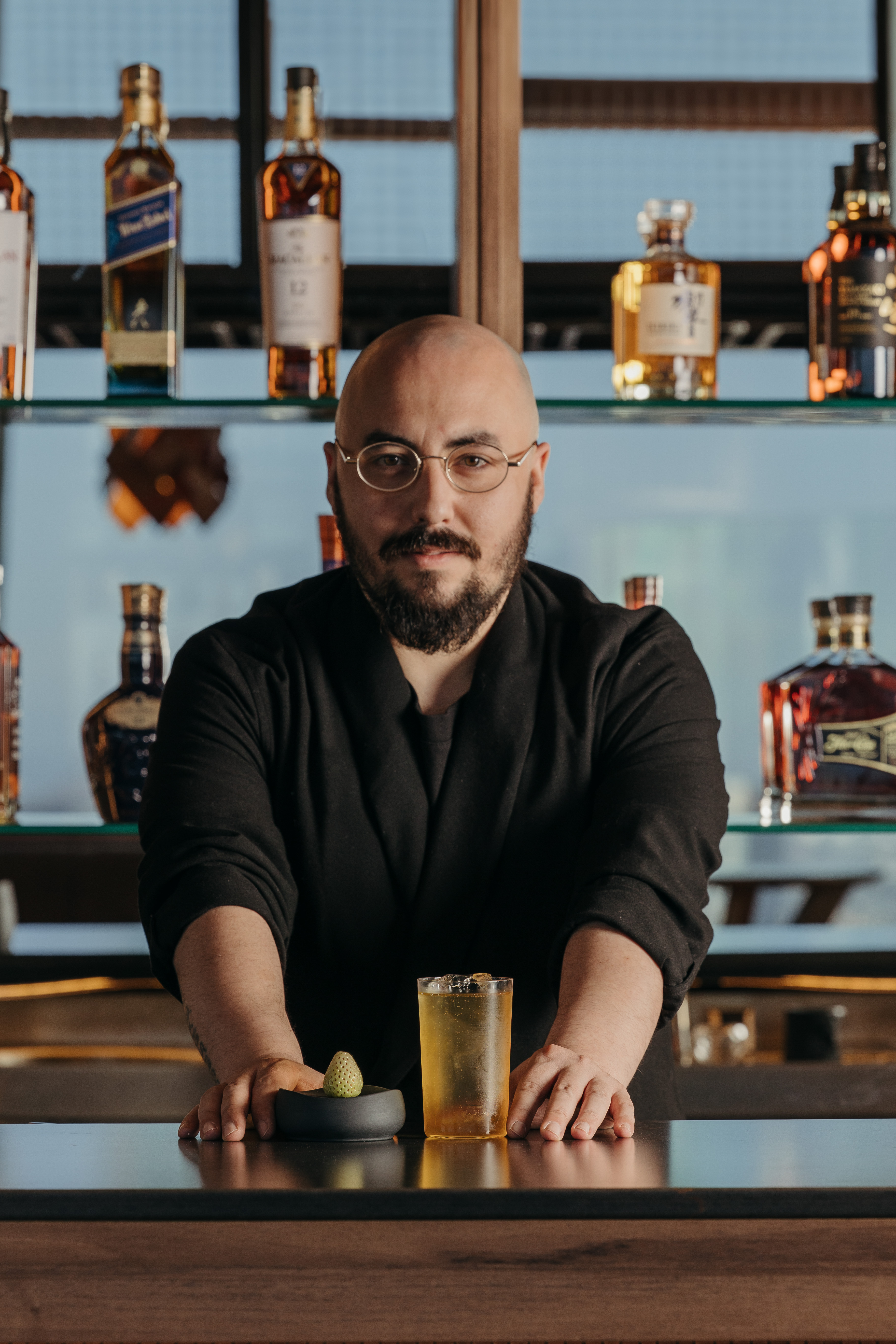 Barman barbudo con gafas presenta cóctel en barra con botellas de whisky.