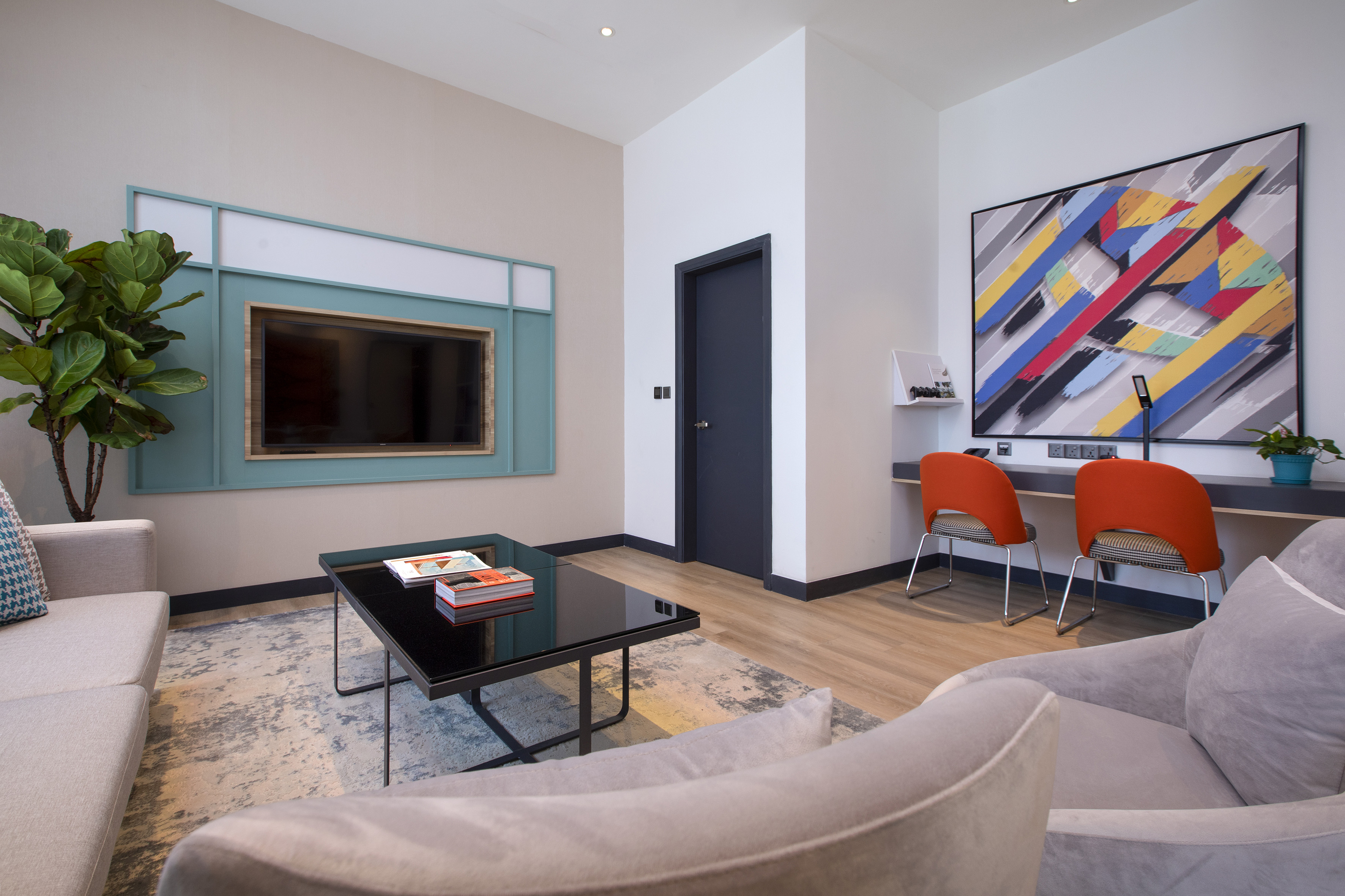 Modern hotel living room with TV, sofa, workspace, orange chairs, and abstract art.