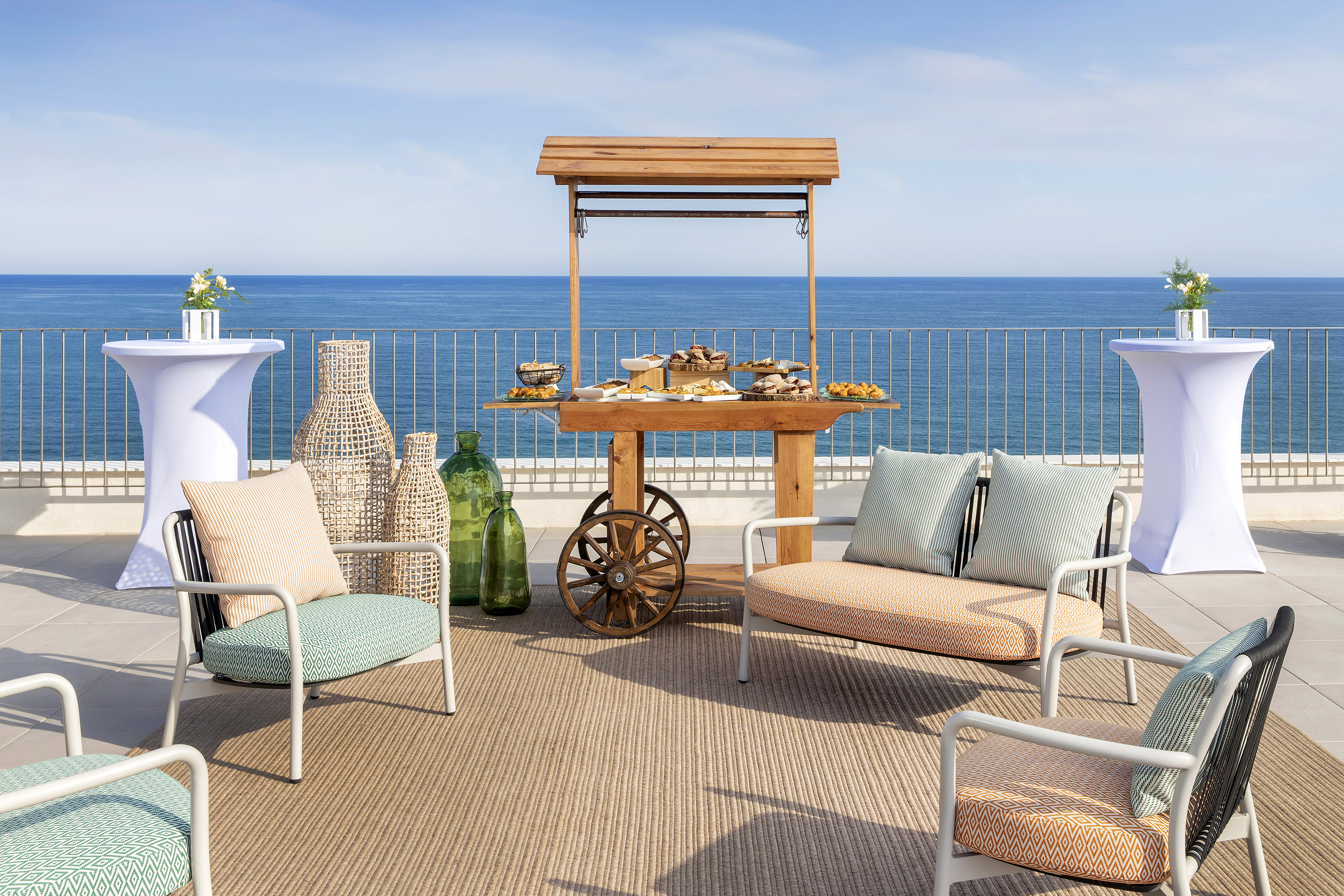 Outdoor terrace with ocean view, buffet cart, lounge seating, and cocktail tables.