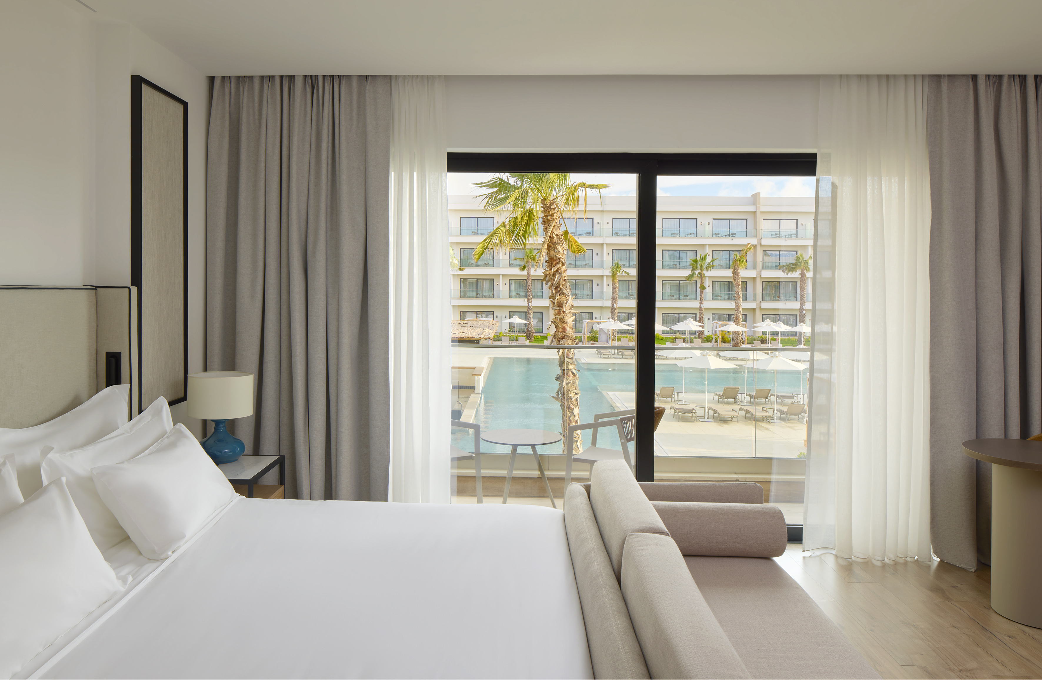 Modern hotel room with balcony overlooking resort pool and palm trees.