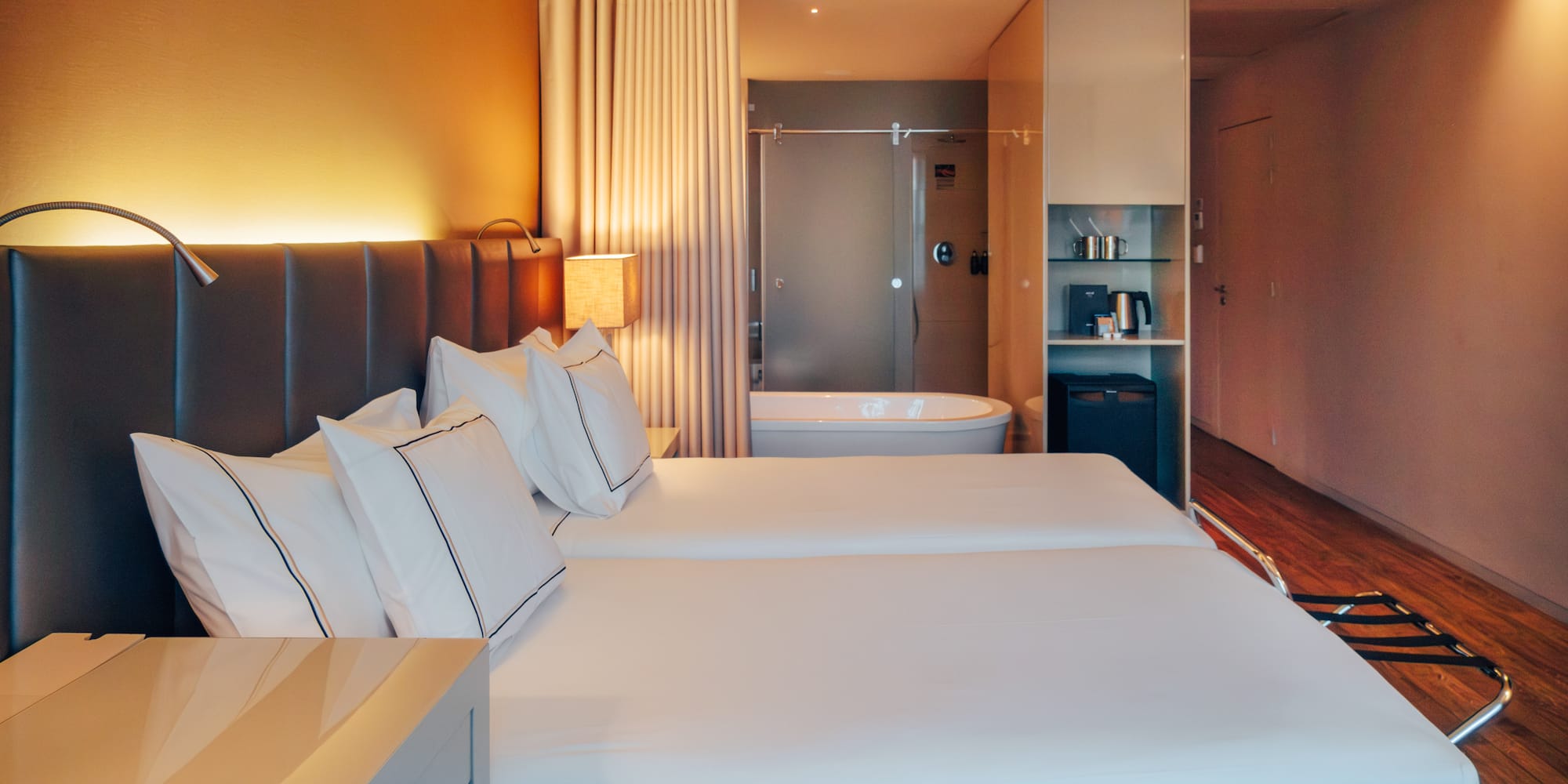 Modern hotel room with twin beds, open-concept bathroom, and warm lighting.