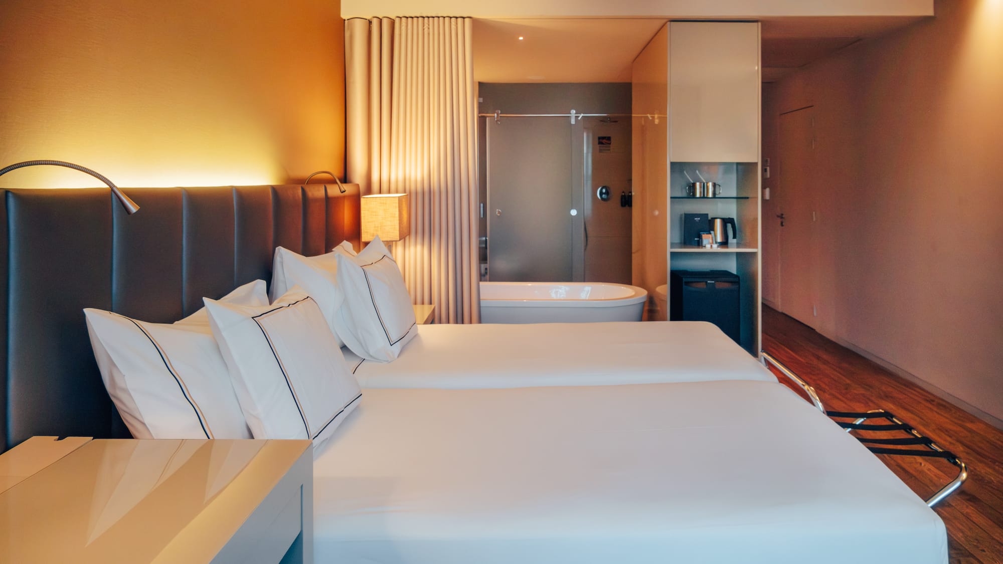 Modern hotel room with twin beds, open-concept bathroom, and warm lighting.
