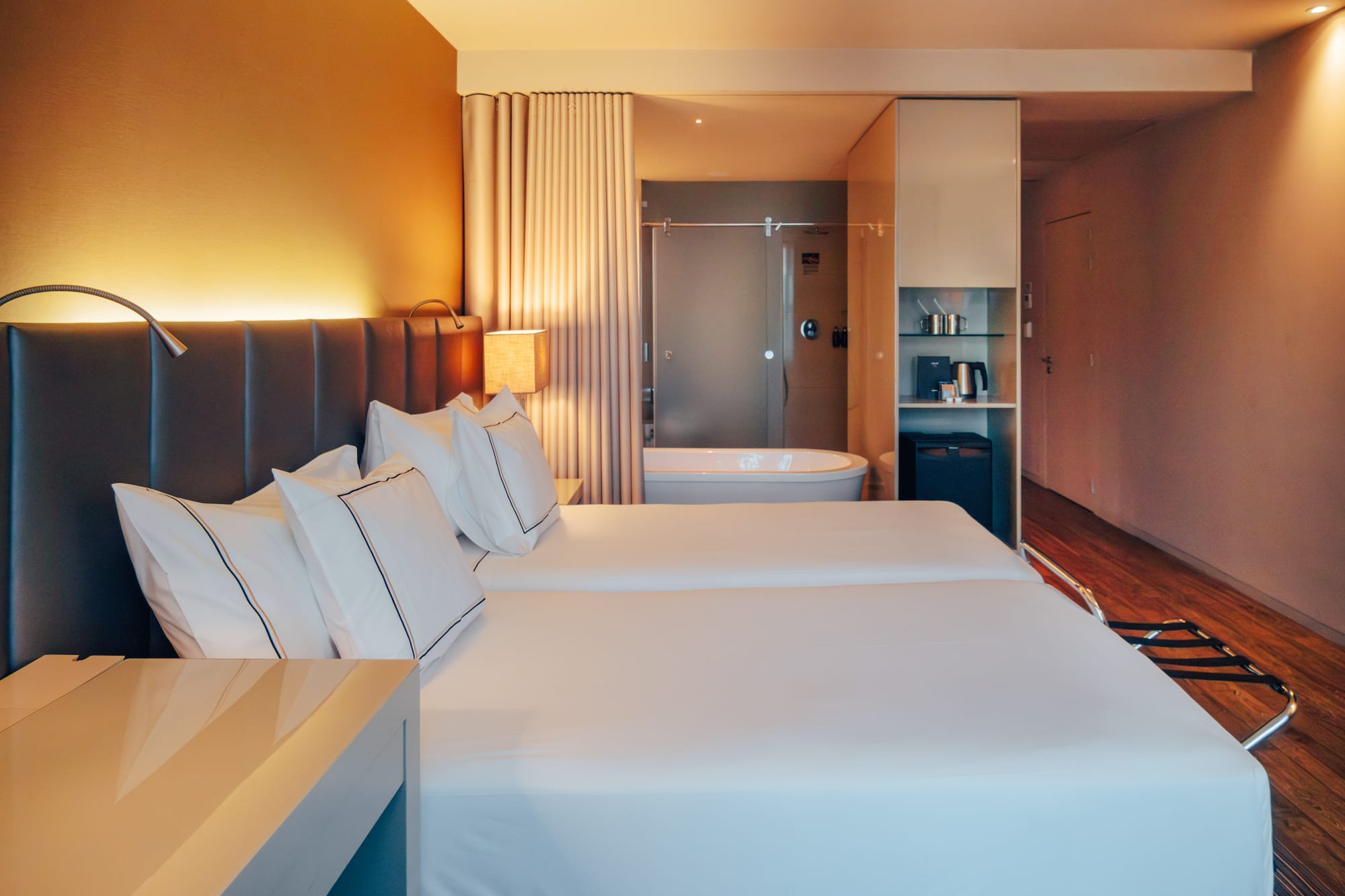Modern hotel room with twin beds, open-concept bathroom, and warm lighting.