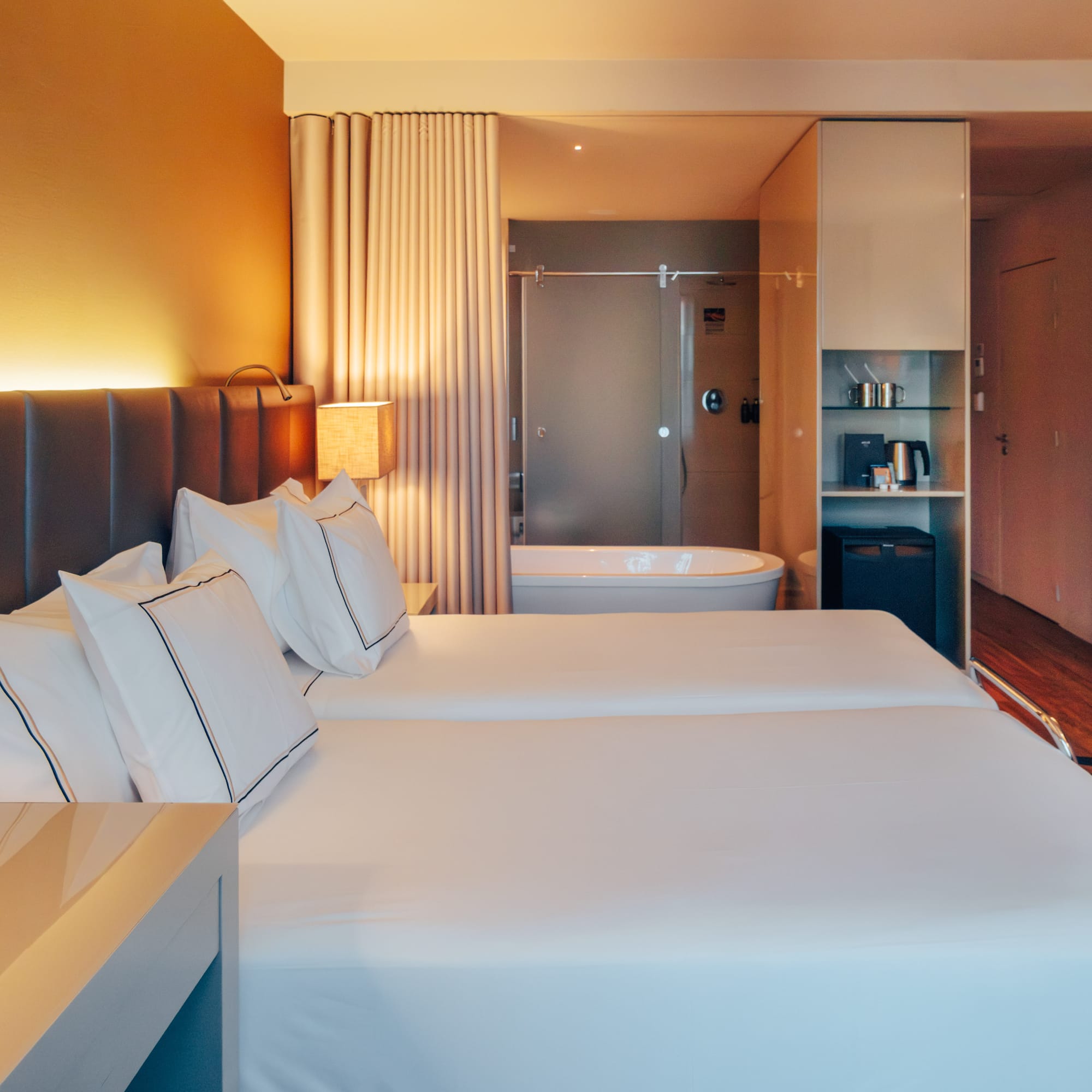 Modern hotel room with twin beds, open-concept bathroom, and warm lighting.
