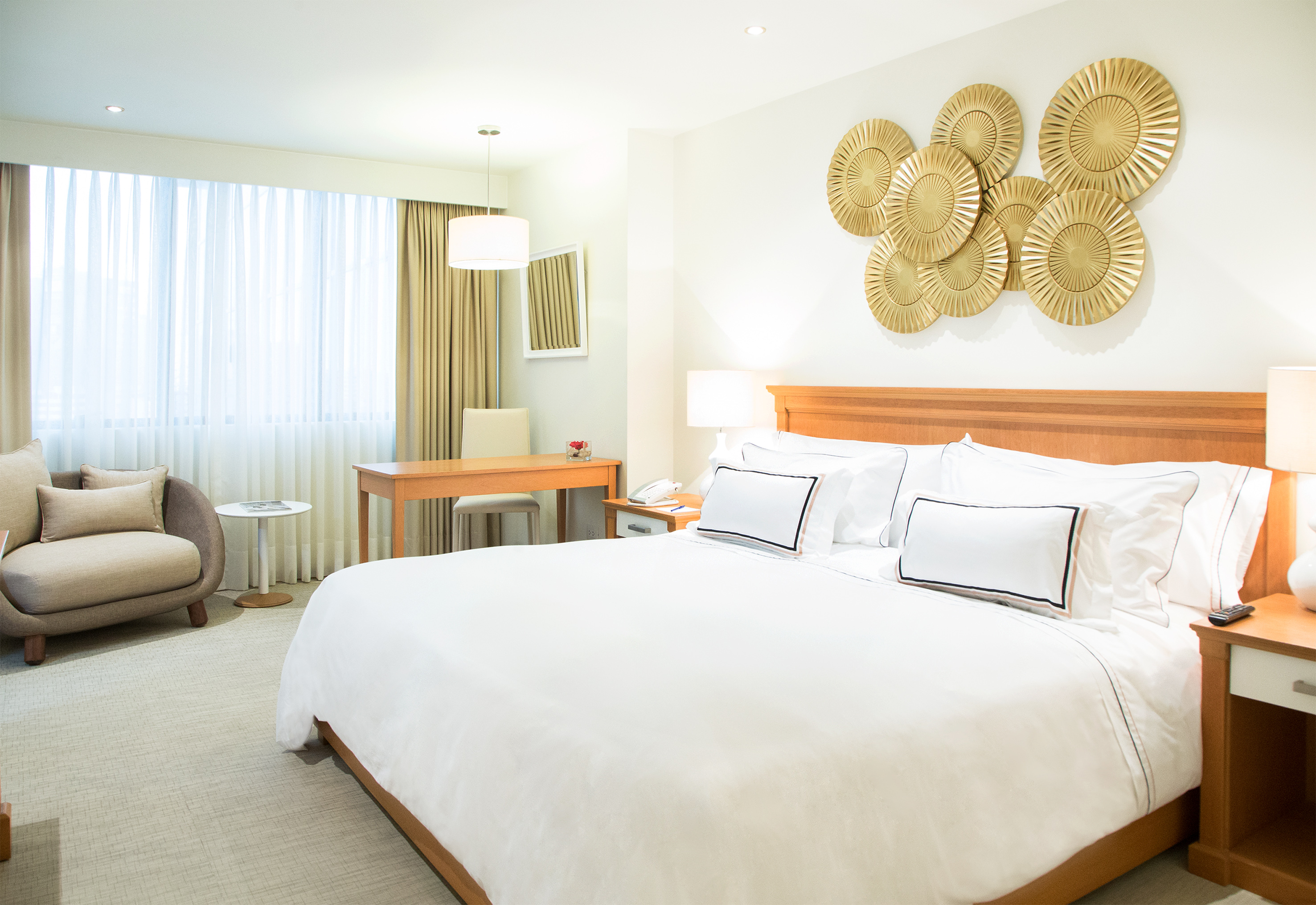 Modern hotel room with king bed, gold wall art, desk, and armchair.