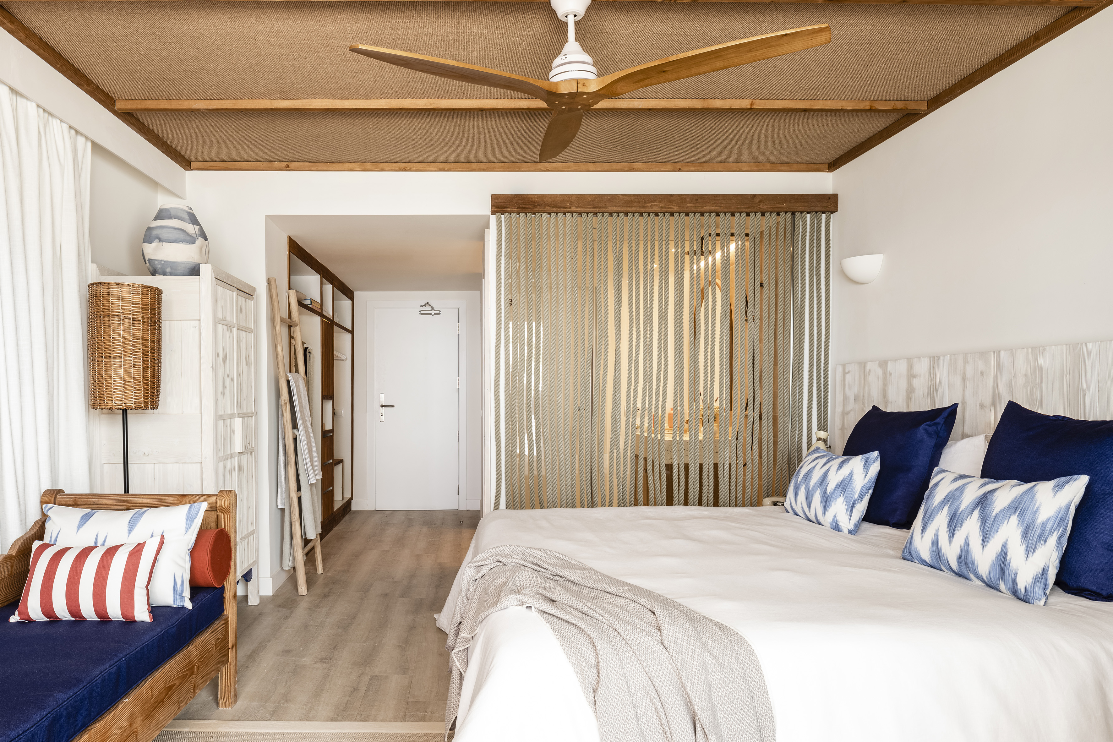 Modern coastal bedroom with large bed, blue pillows, wooden ceiling fan, and daybed.