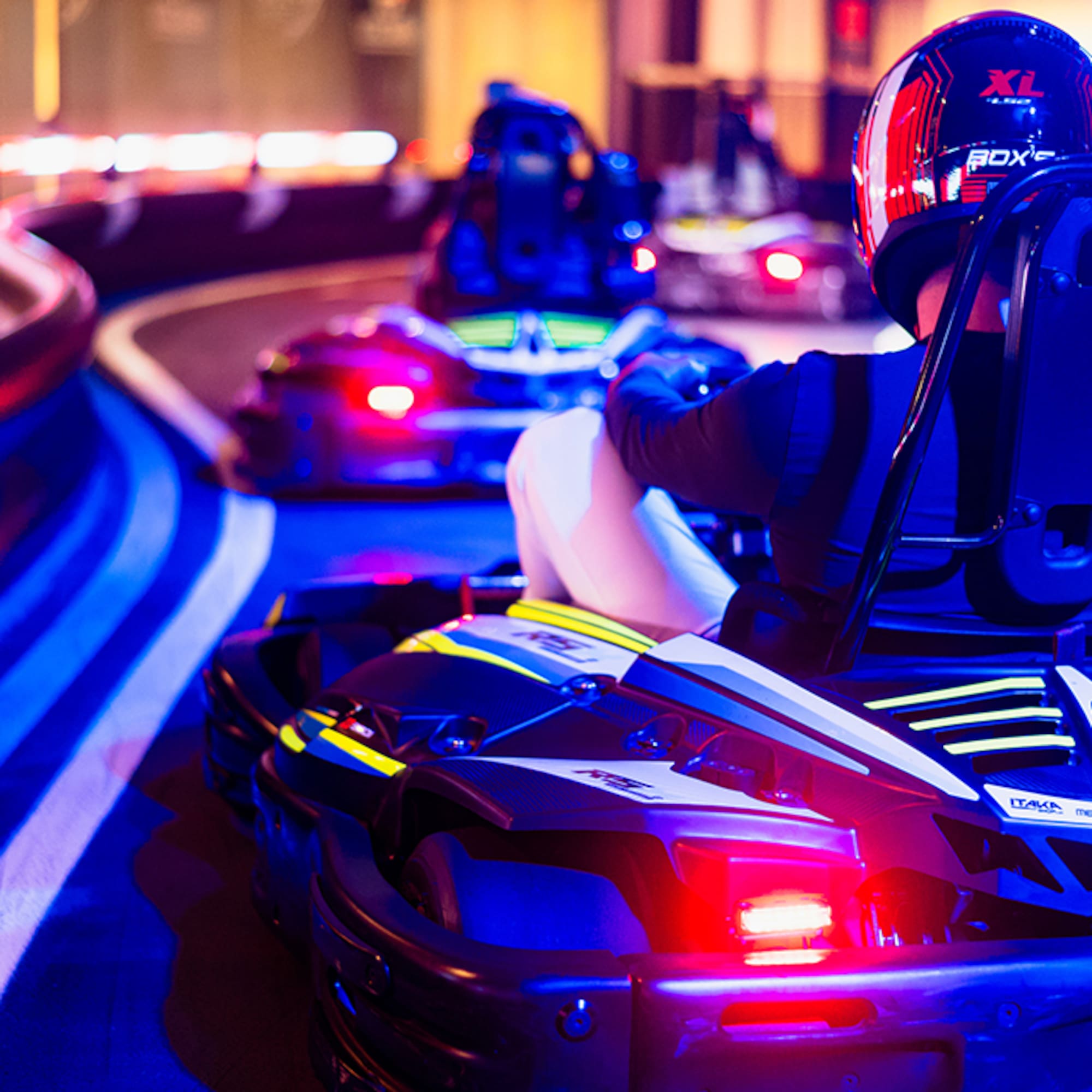 Driver in a helmet on a go-kart, racing on a neon-lit indoor track.