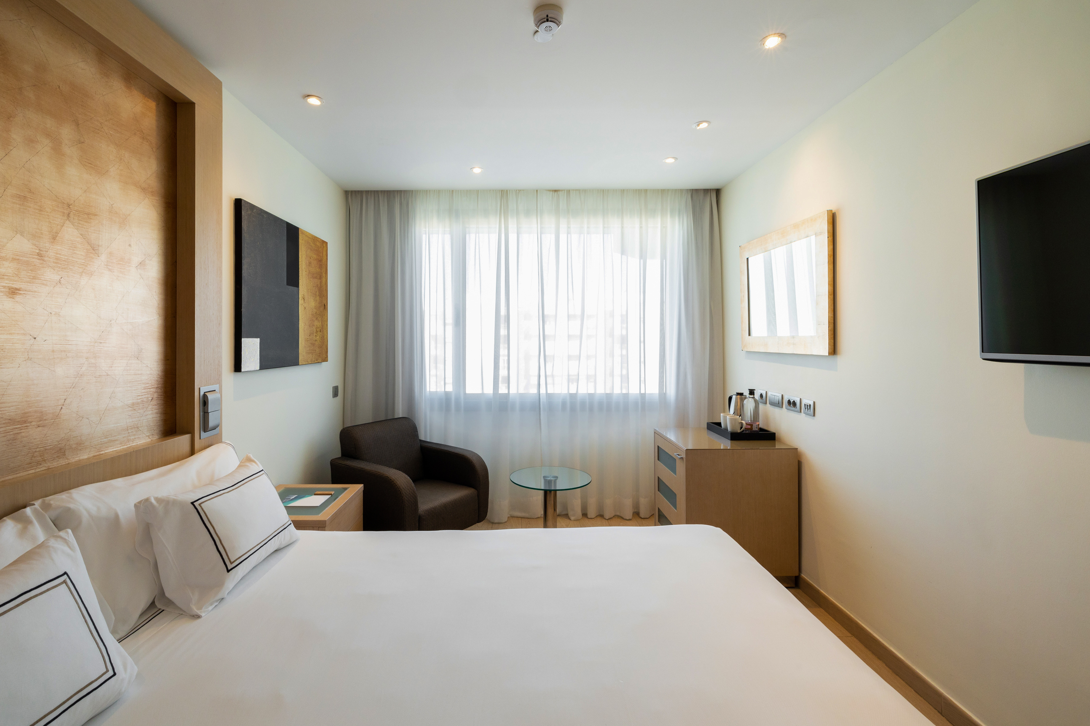 Contemporary hotel room with bed, armchair, TV, and sheer window curtains.