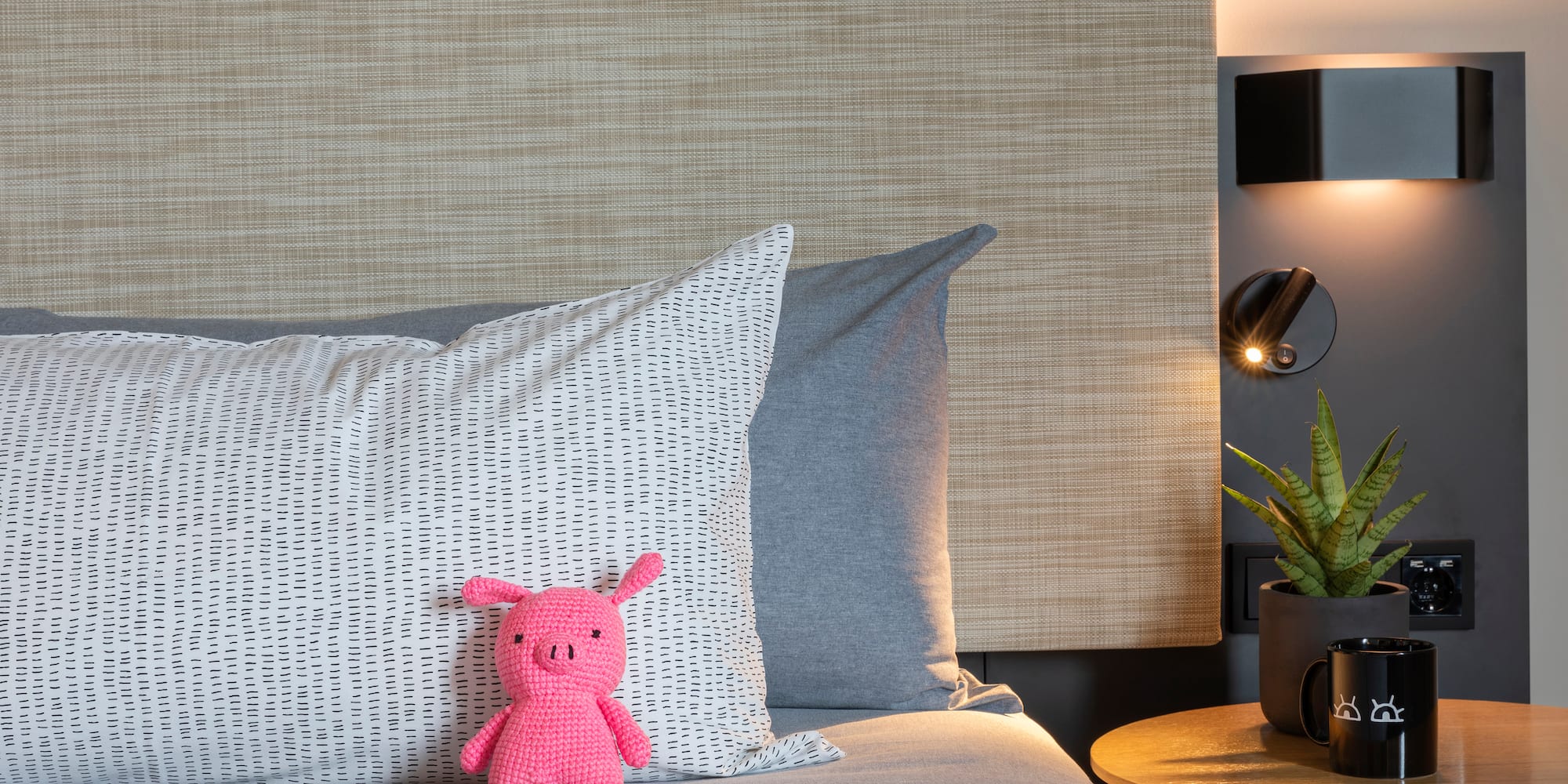 Pink knitted pig toy on modern bed with patterned pillow, bedside lamp, and plant.