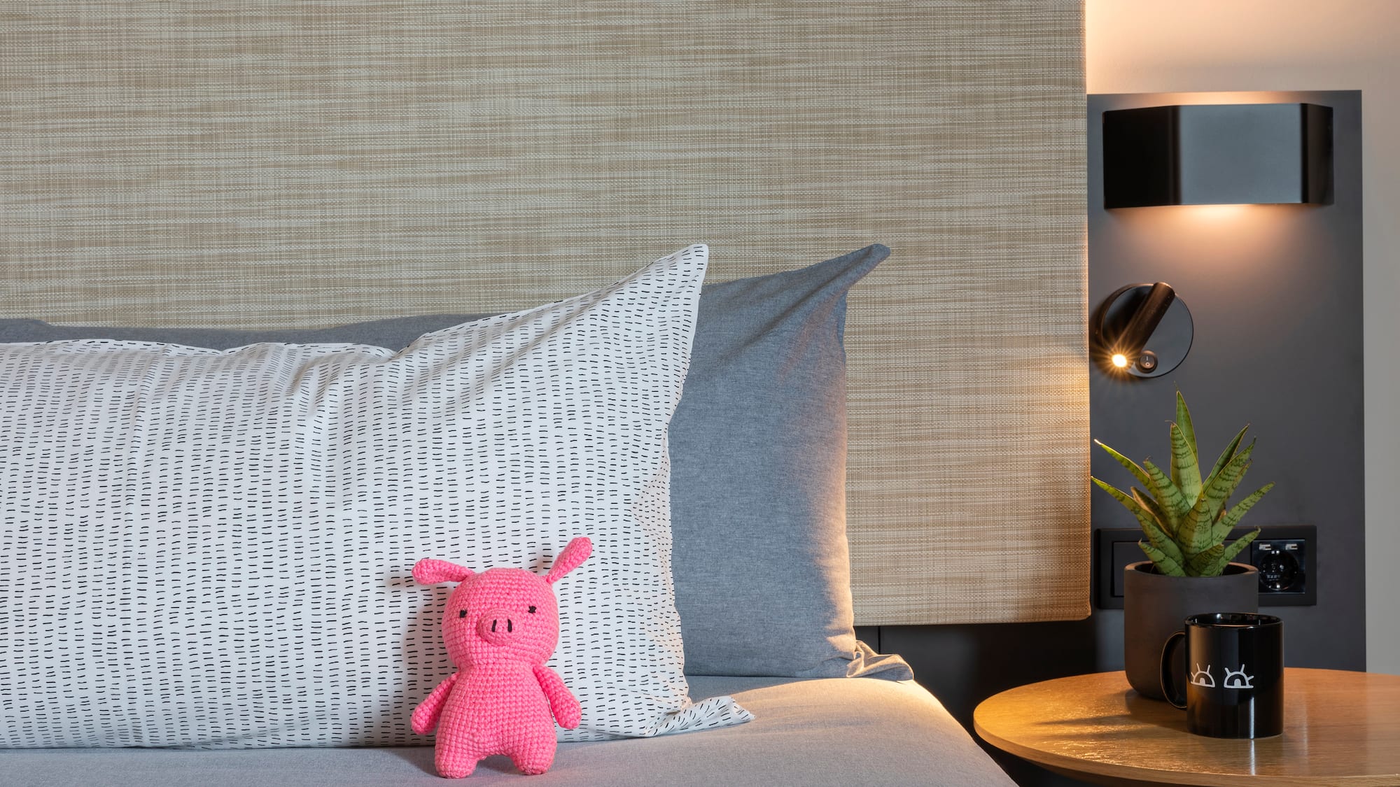 Pink crochet pig on bed, patterned pillow, nightstand with plant and warm lamp.