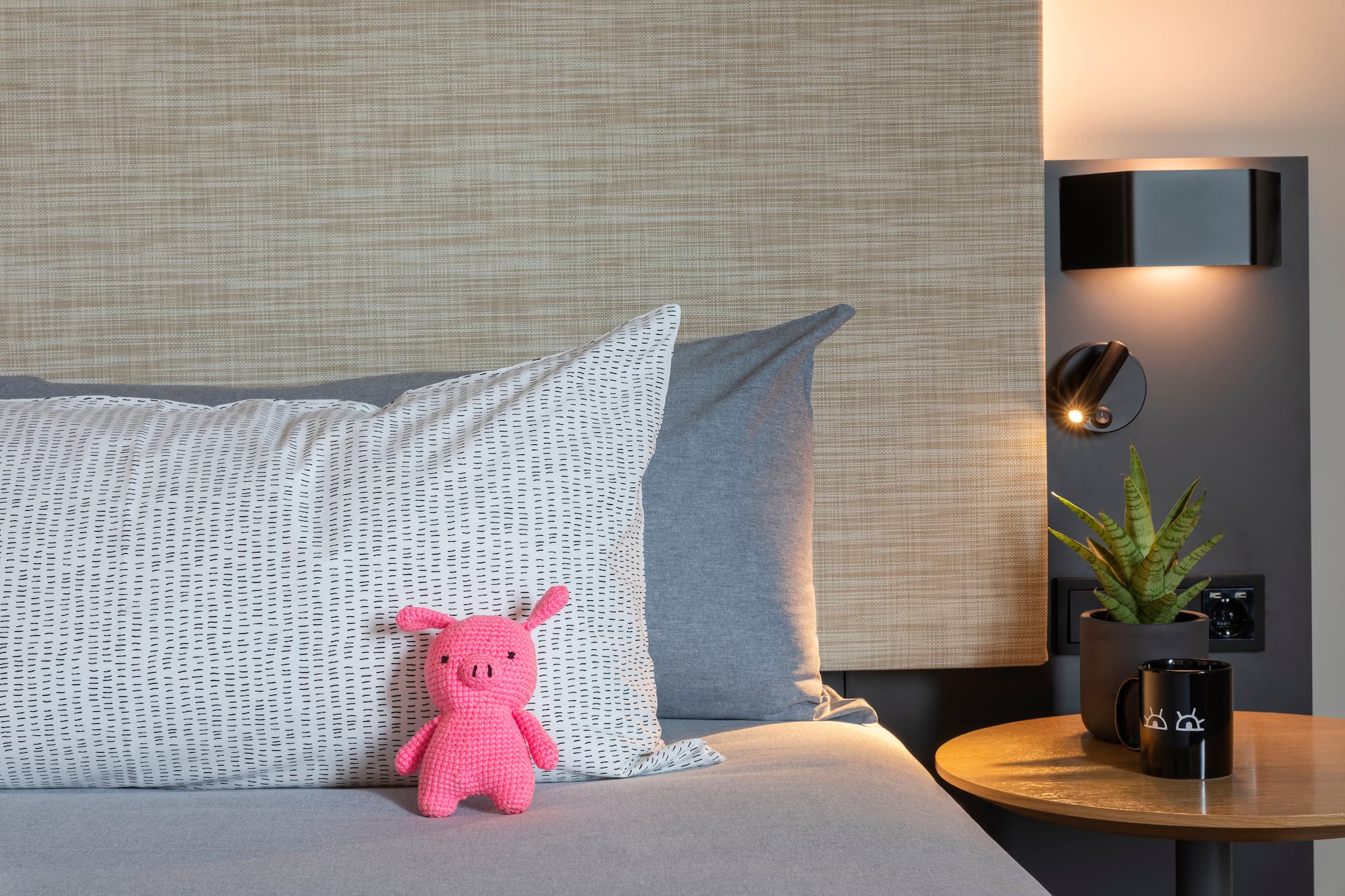 Pink crochet pig on bed, patterned pillow, nightstand with plant and warm lamp.