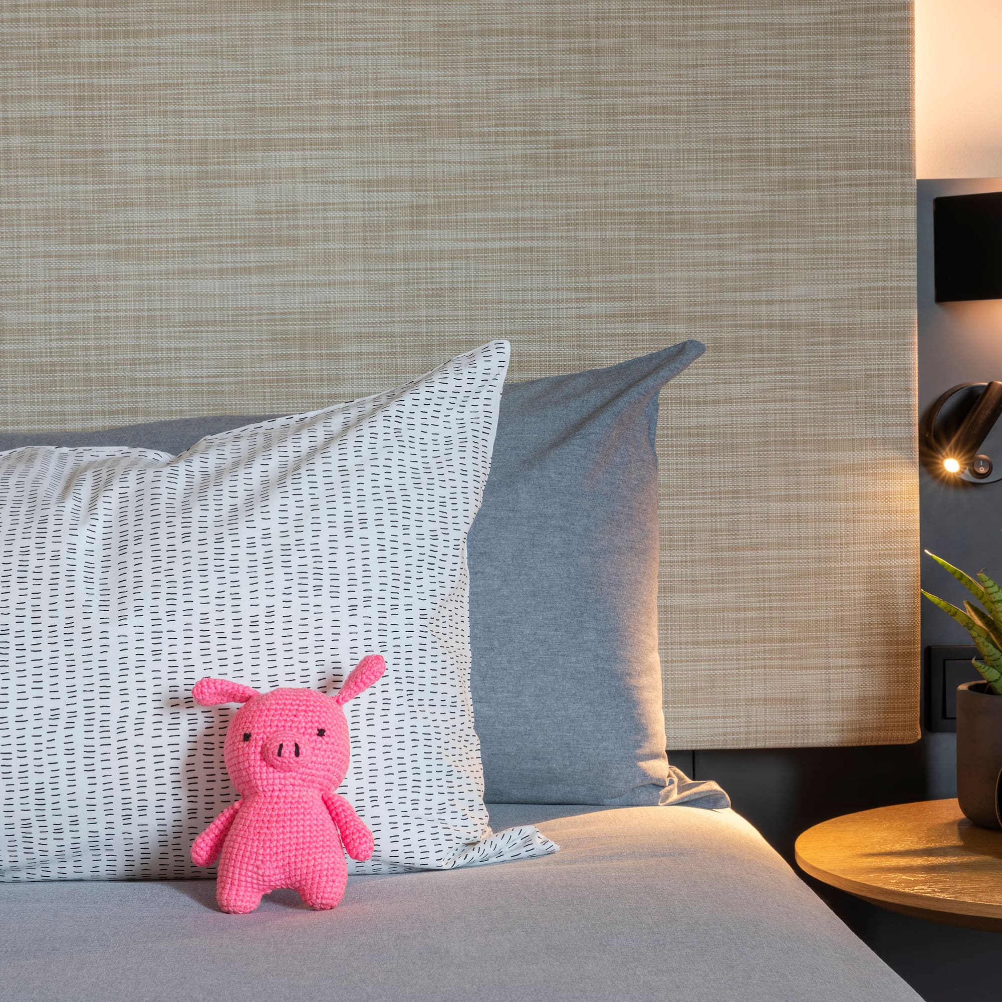 Pink crochet pig on bed, patterned pillow, nightstand with plant and warm lamp.