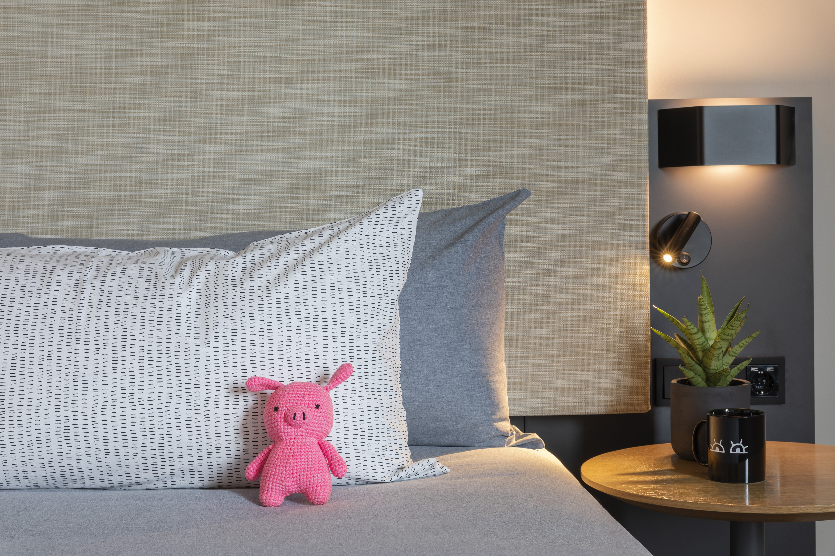 Pink knitted pig toy on modern bed with patterned pillow, bedside lamp, and plant.