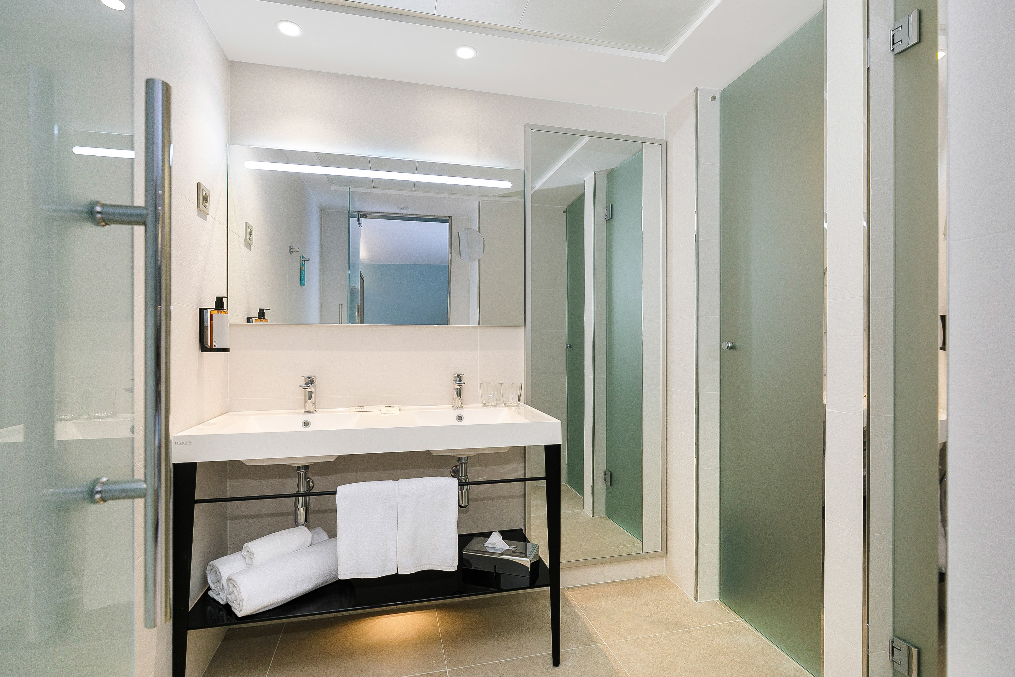 Sleek hotel bathroom with double vanity, illuminated mirror, and glass partitions.