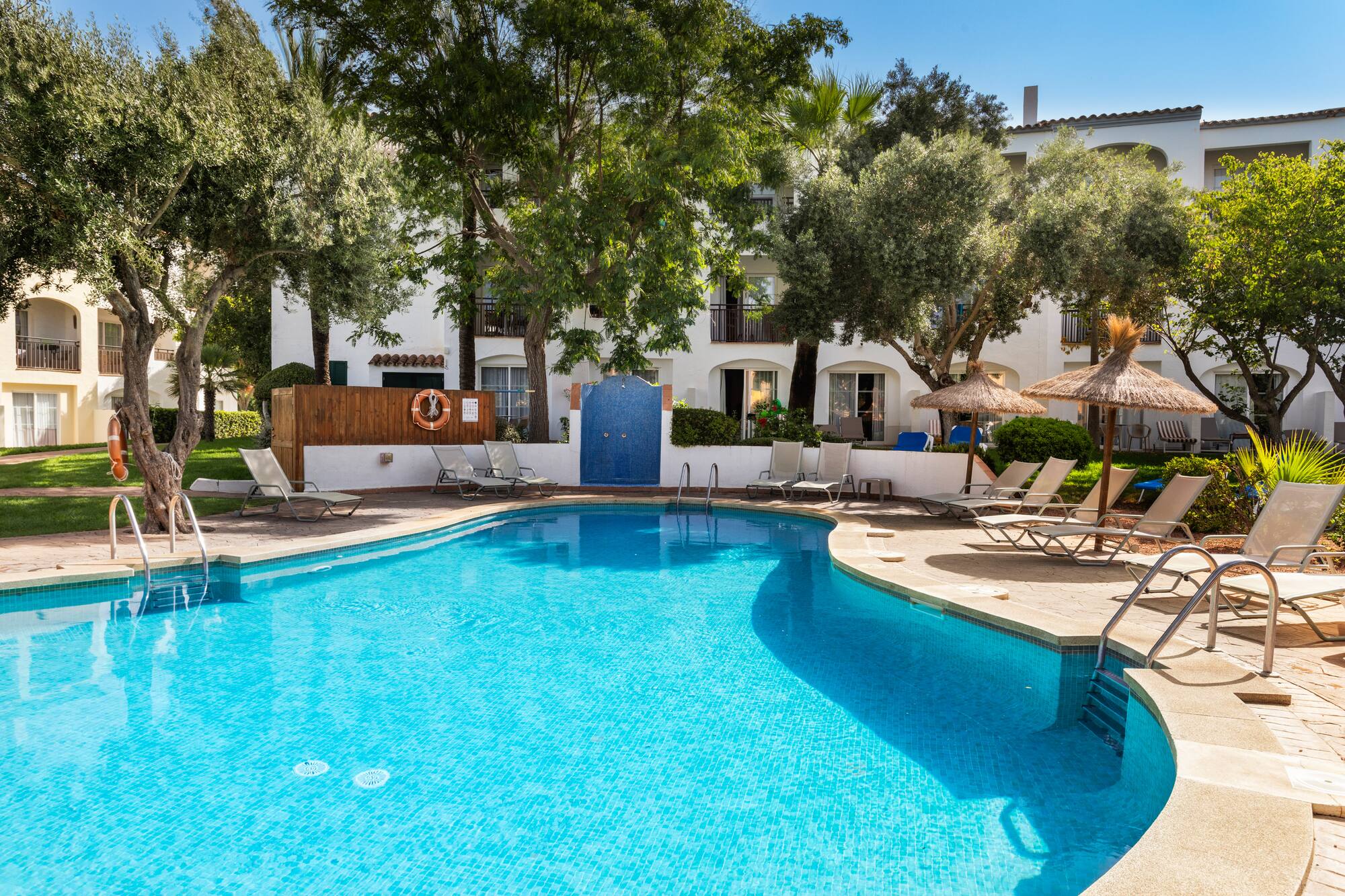 Sunny resort pool with lounge chairs, straw umbrellas, and hotel buildings.