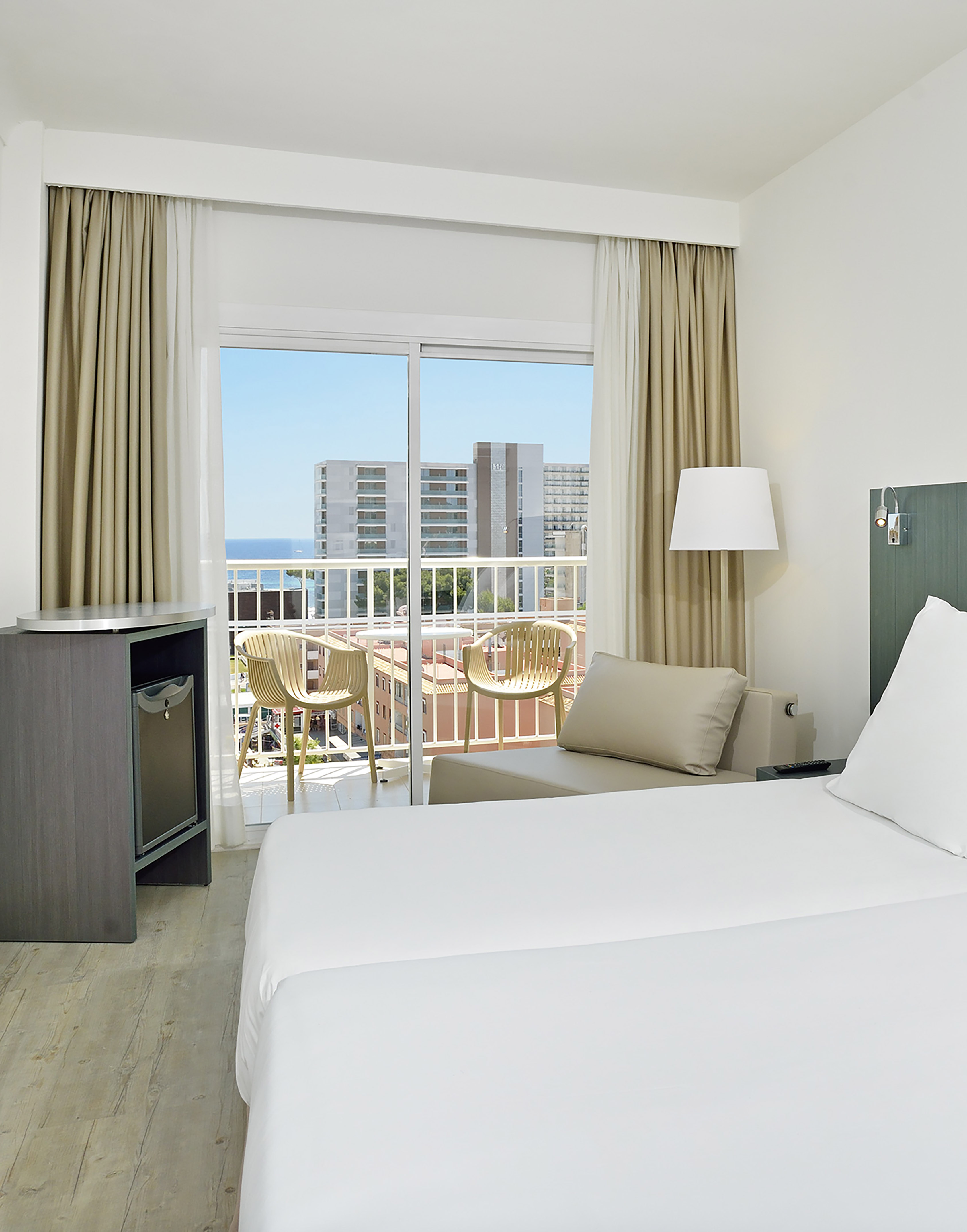 Bright hotel room with sea view balcony, bed, and outdoor seating.