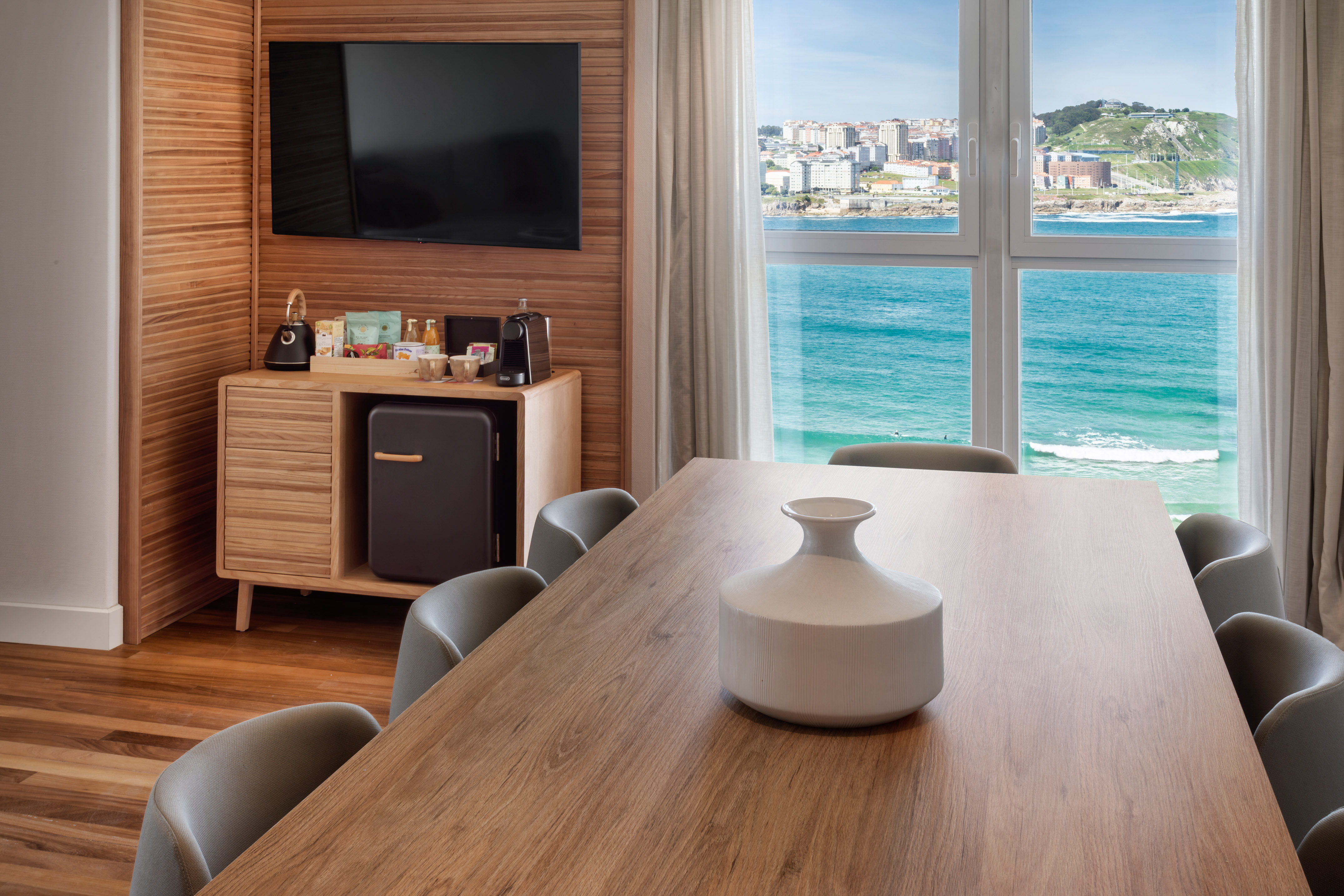 Modern hotel room with ocean view, wooden dining table, and sleek mini-bar.