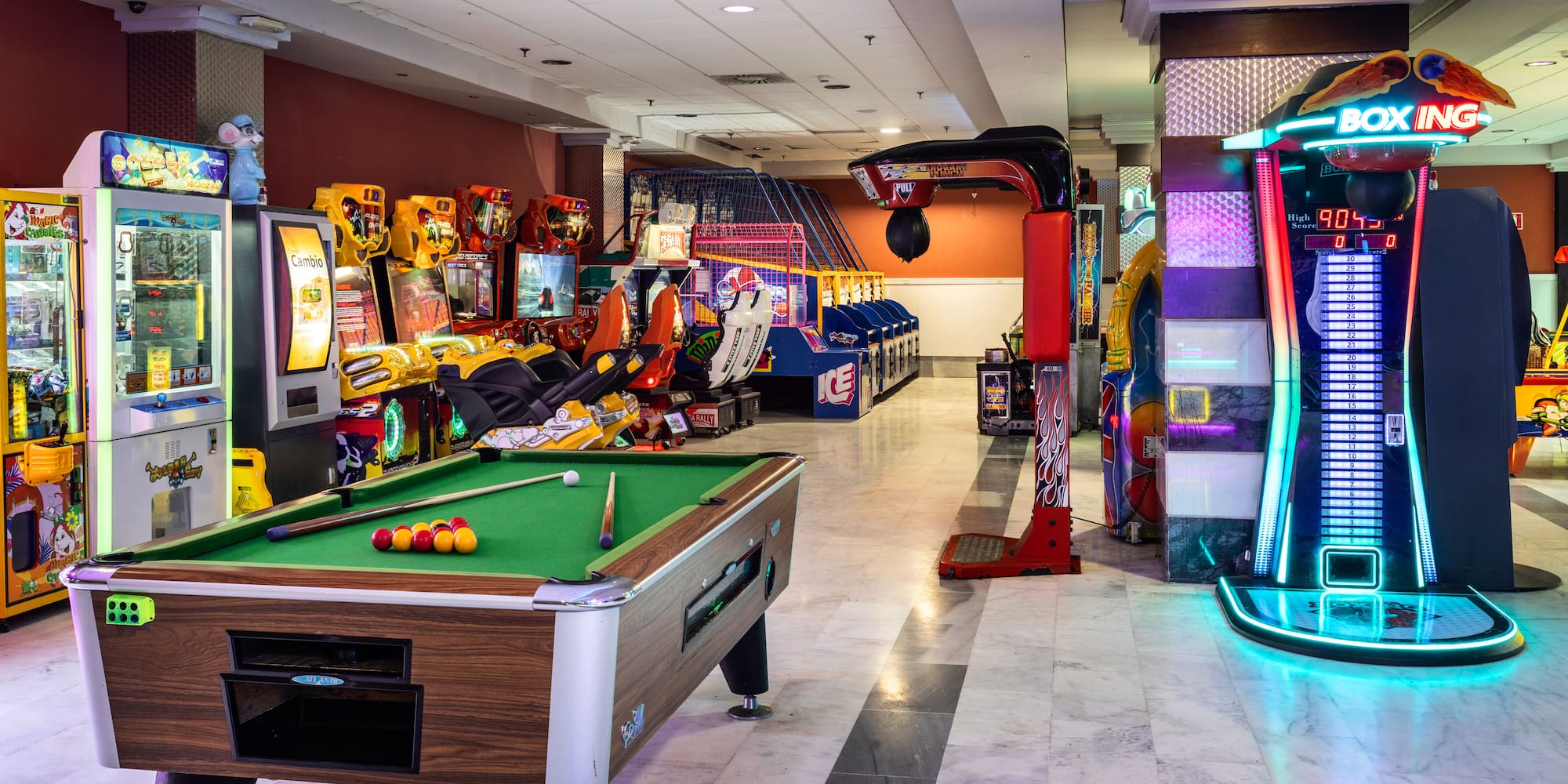 Brightly lit arcade game room with pool table, racing games, and boxing machine.