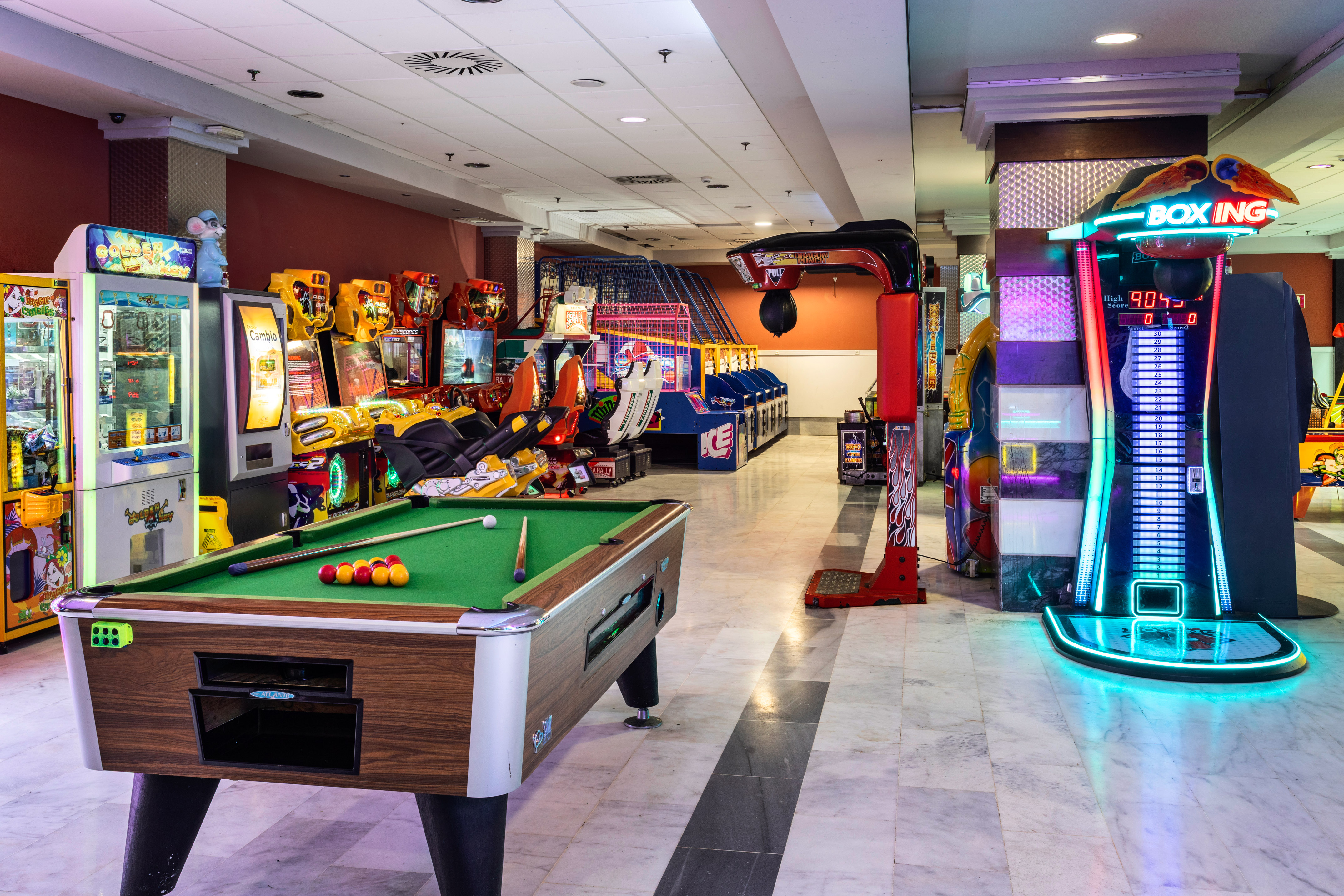 Brightly lit arcade game room with pool table, racing games, and boxing machine.