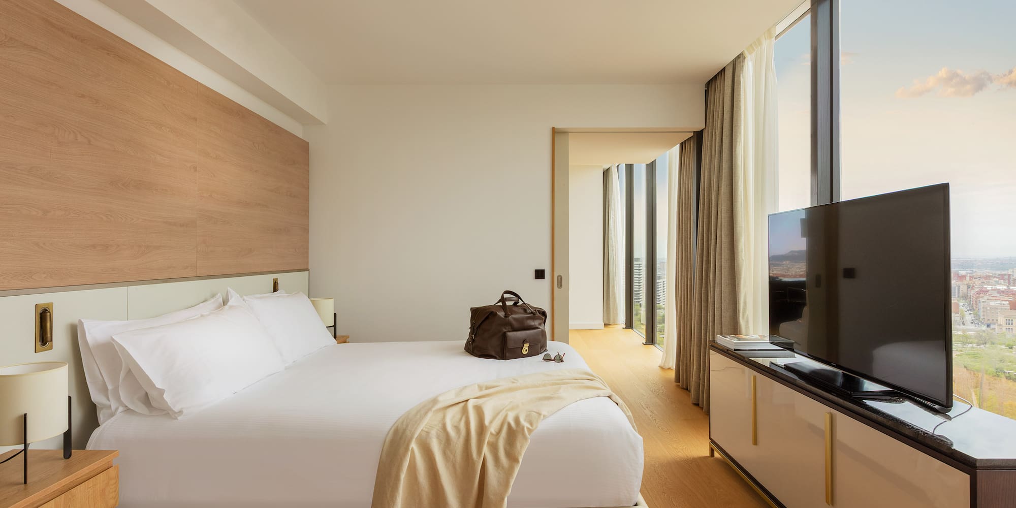 Modern hotel room with white bed, floor-to-ceiling city view windows.