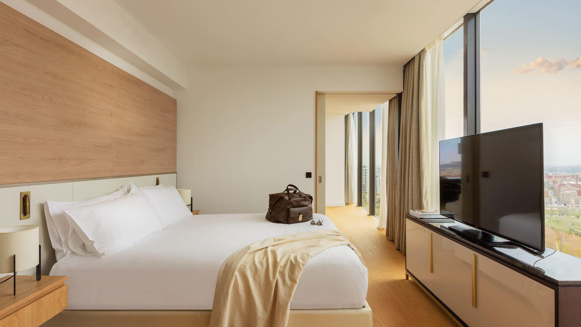 Modern hotel room with white bed, floor-to-ceiling city view windows.