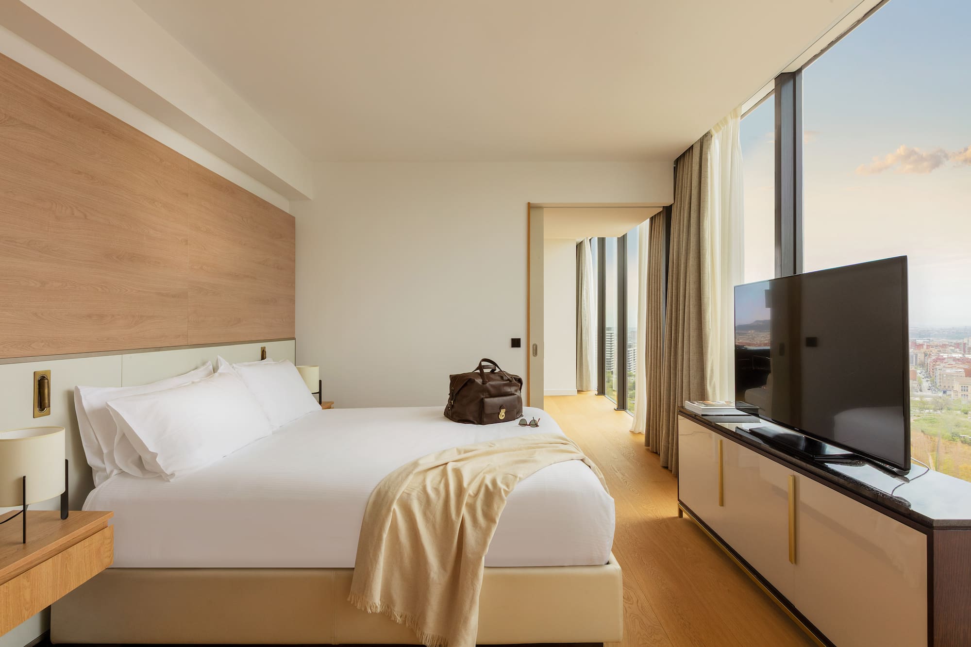 Modern hotel room with white bed, floor-to-ceiling city view windows.
