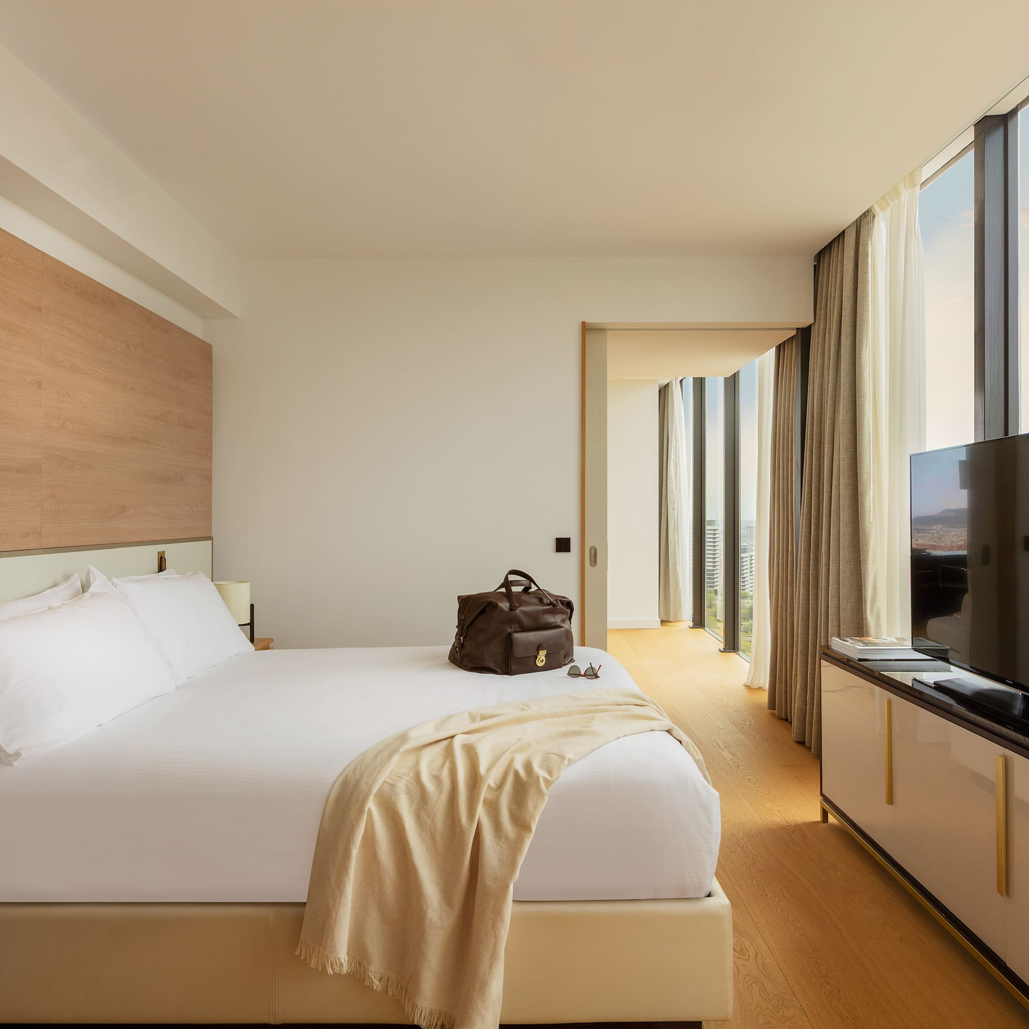 Modern hotel room with white bed, floor-to-ceiling city view windows.