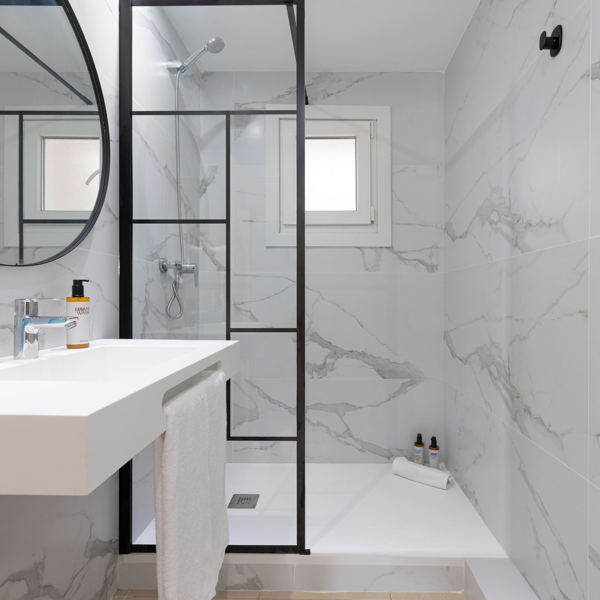 White marble bathroom with glass shower, floating vanity, and round mirror.