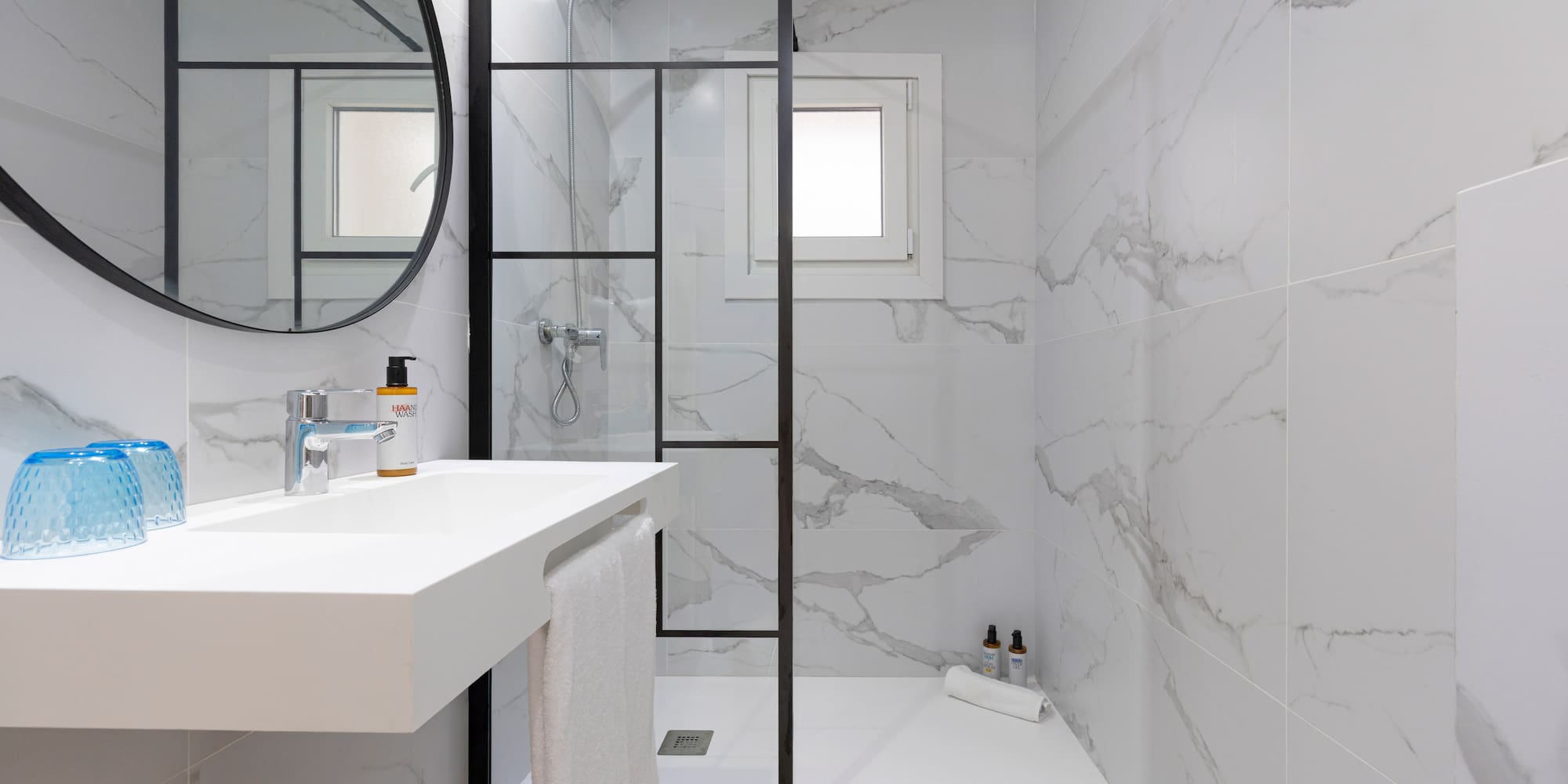 White marble bathroom with glass shower, floating vanity, and round mirror.