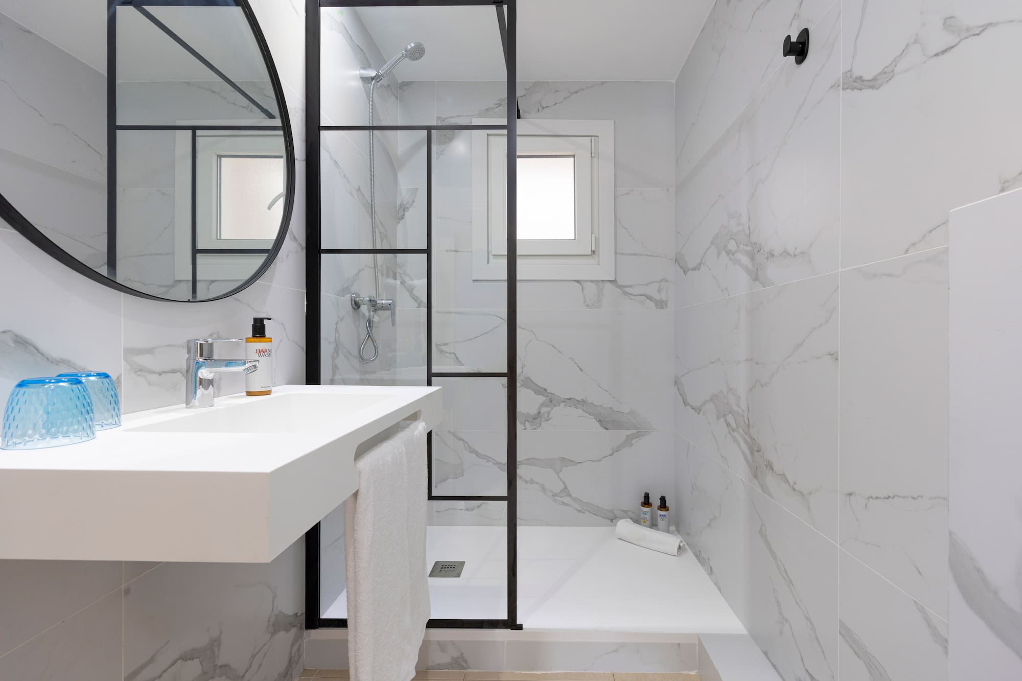 White marble bathroom with glass shower, floating vanity, and round mirror.