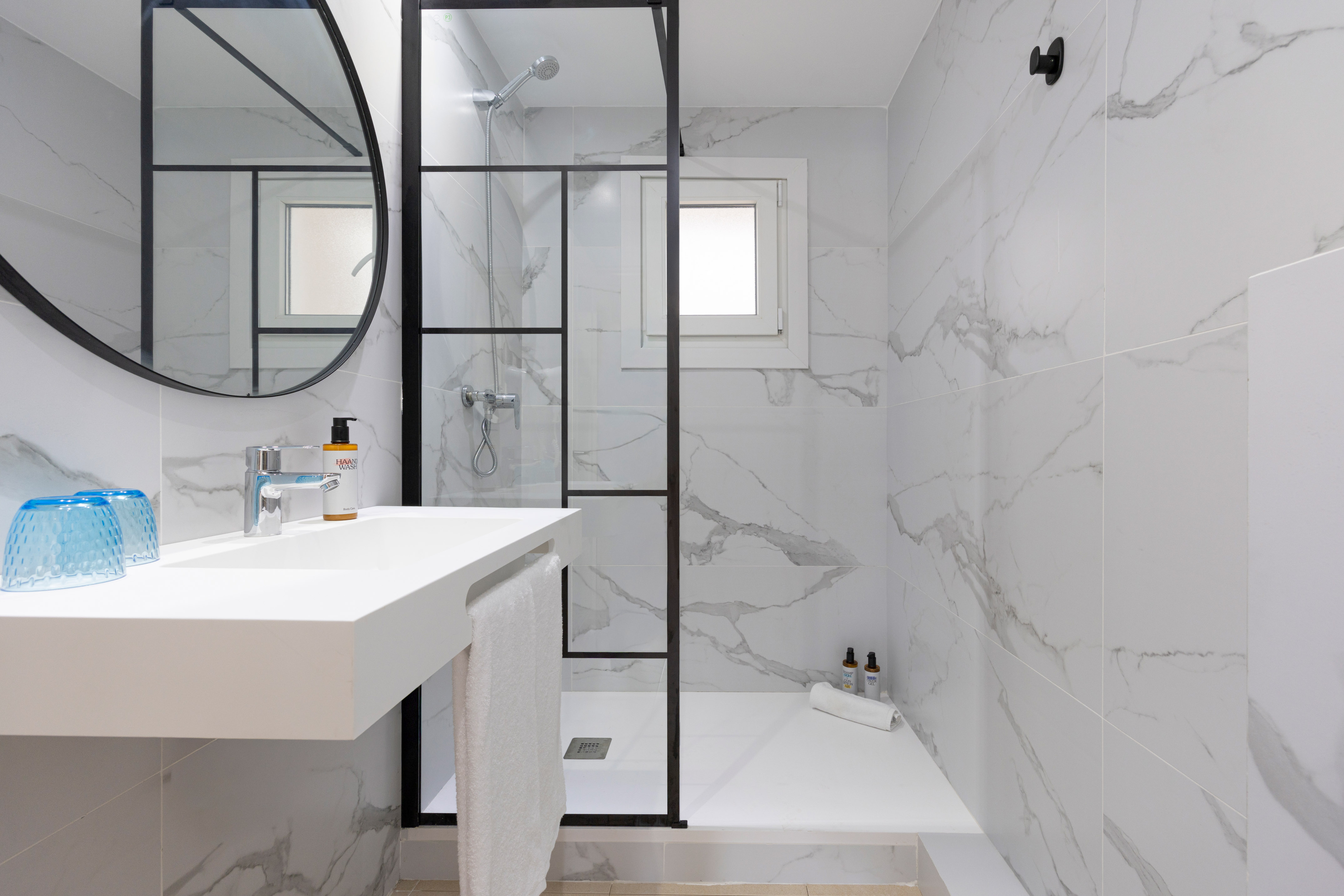White marble bathroom with glass shower, floating vanity, and round mirror.