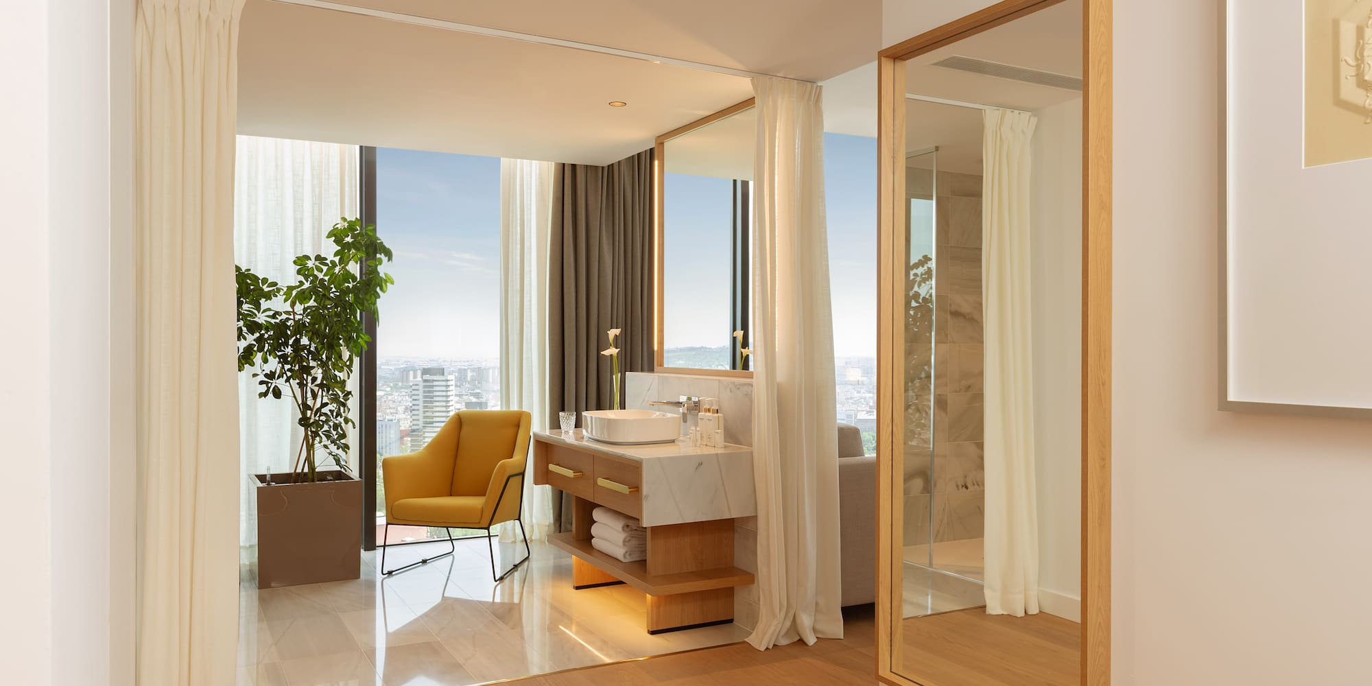 Luxury hotel suite with modern bathroom vanity, city view, and yellow armchair.