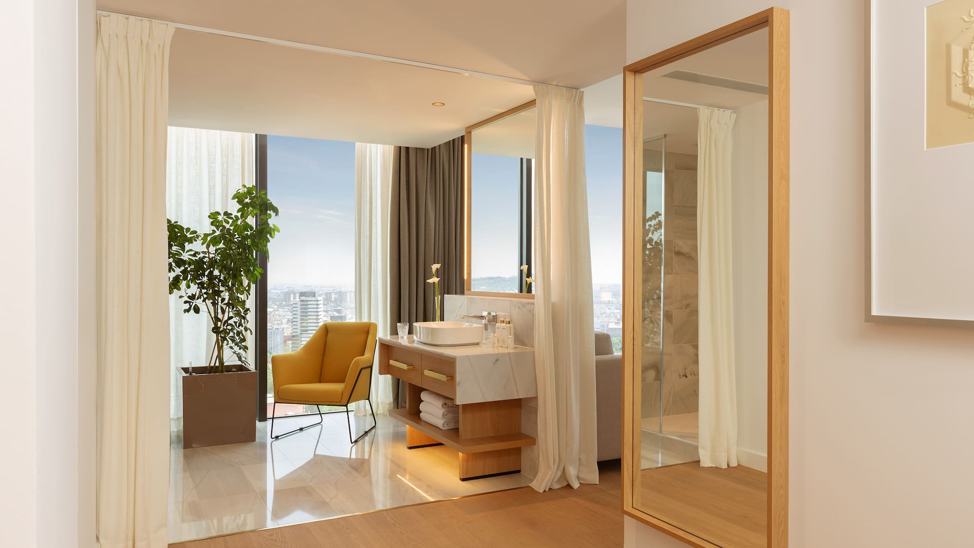 Luxury hotel suite with modern bathroom vanity, city view, and yellow armchair.