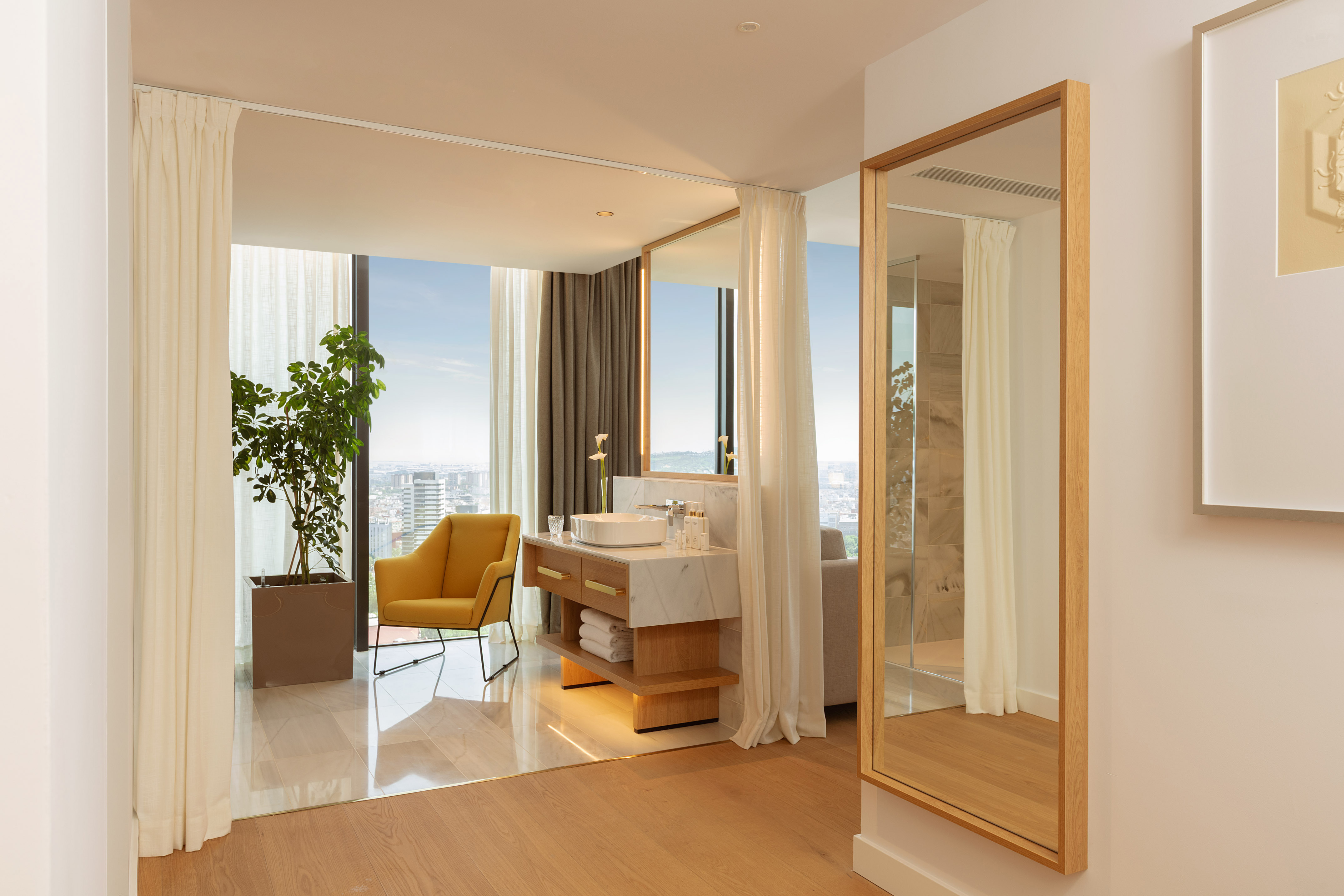 Luxury hotel suite with modern bathroom vanity, city view, and yellow armchair.