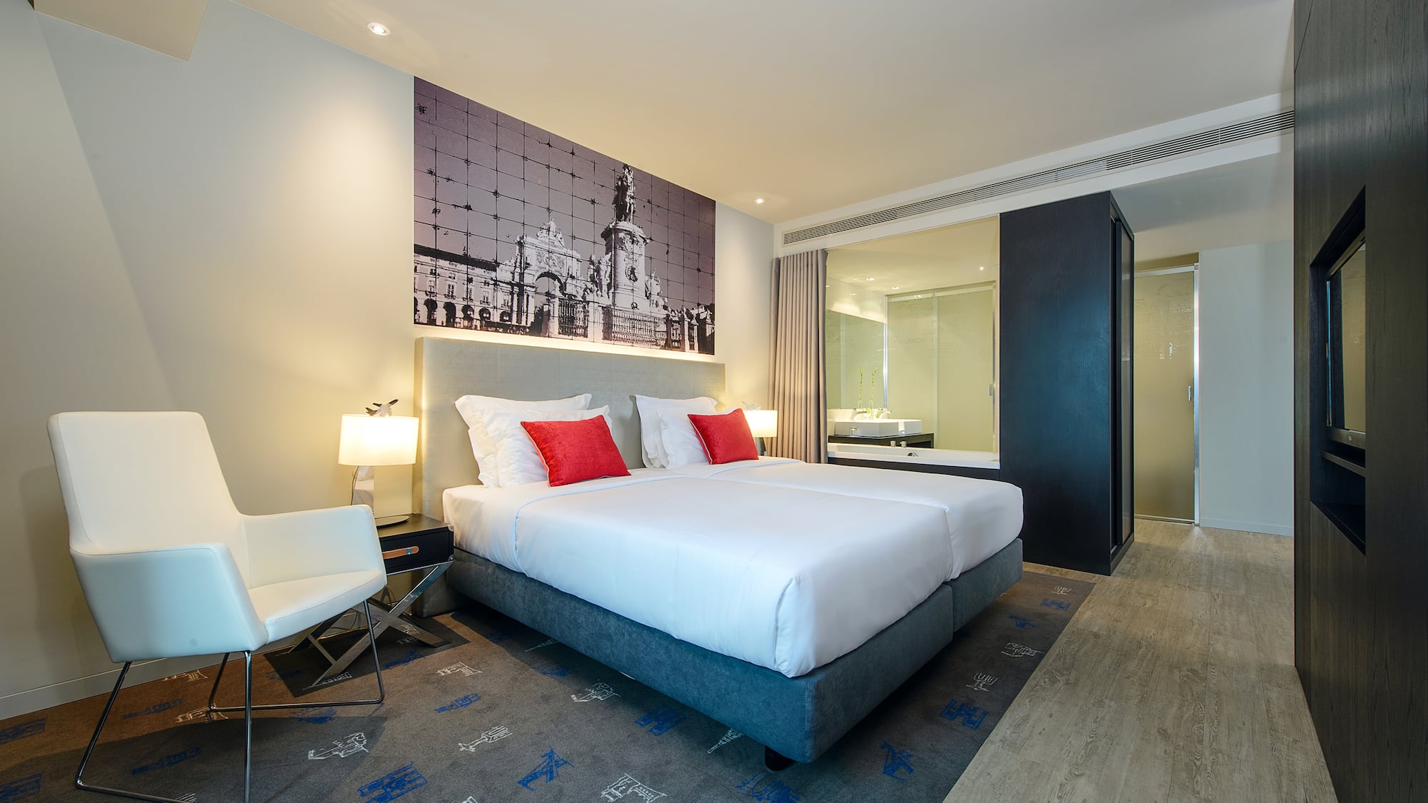 Lisbon hotel room with bed, red pillows, city arch mural, and glass bathroom.