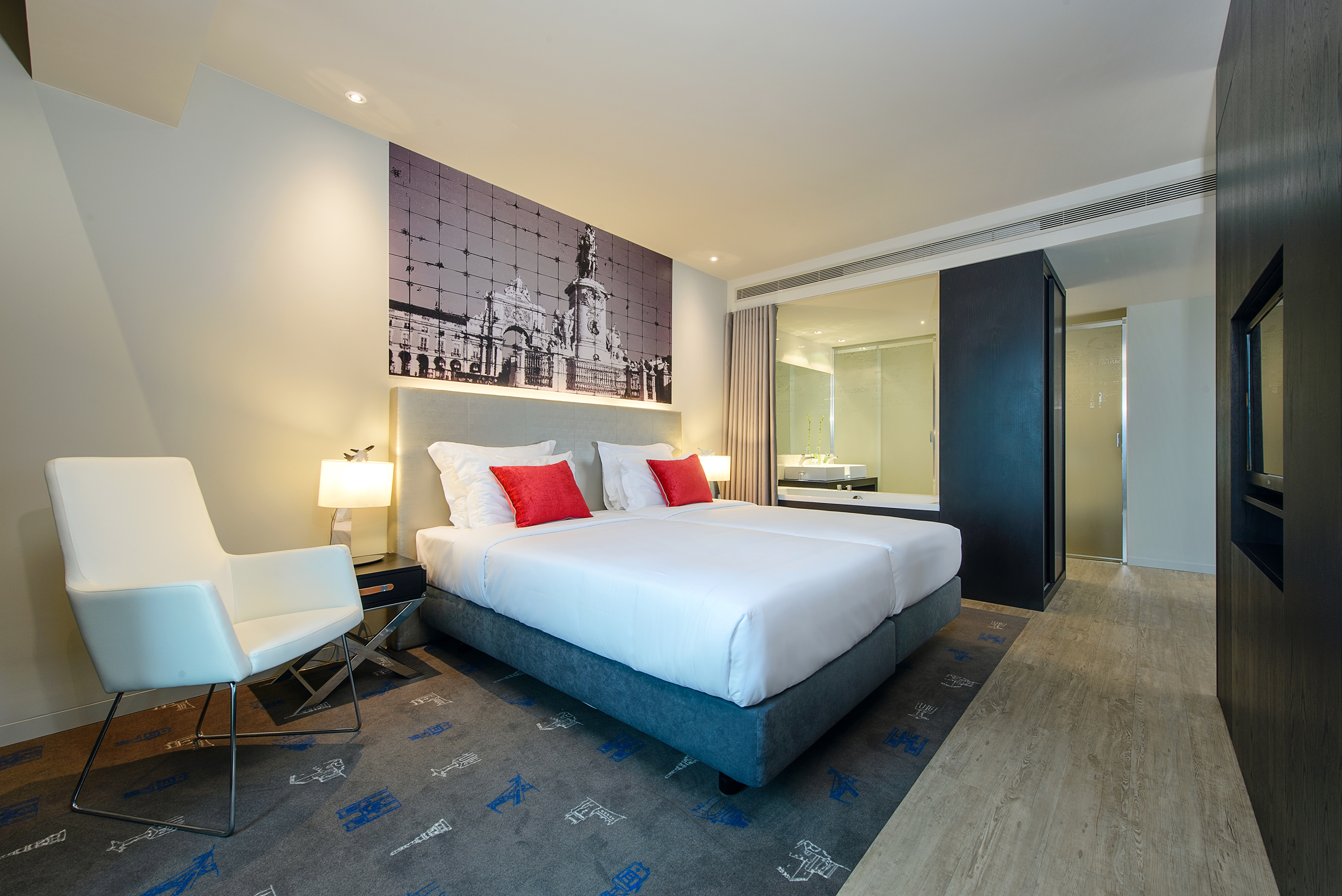 Lisbon hotel room with bed, red pillows, city arch mural, and glass bathroom.