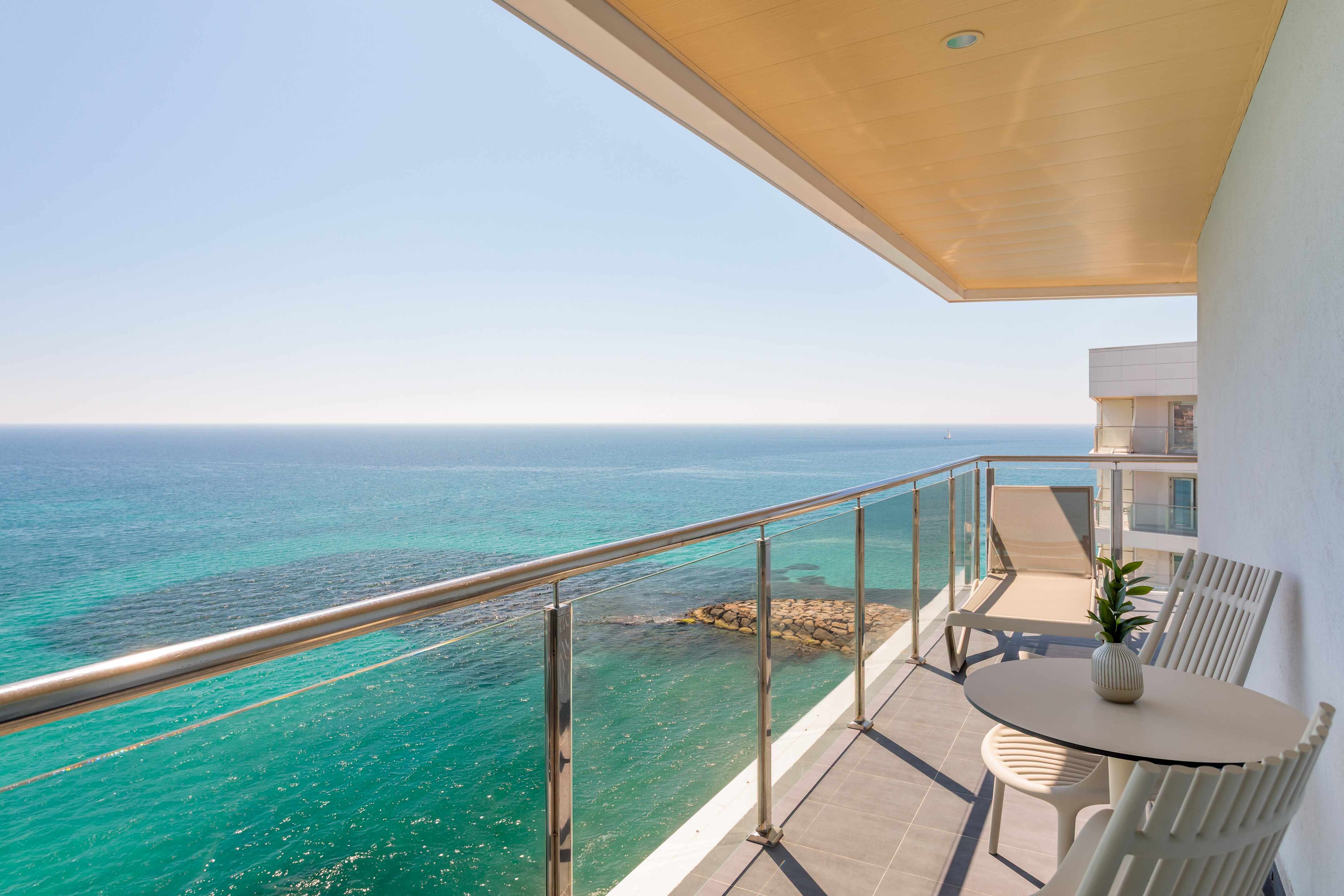 Hotel balcony with turquoise sea view, lounge chair, and outdoor furniture.