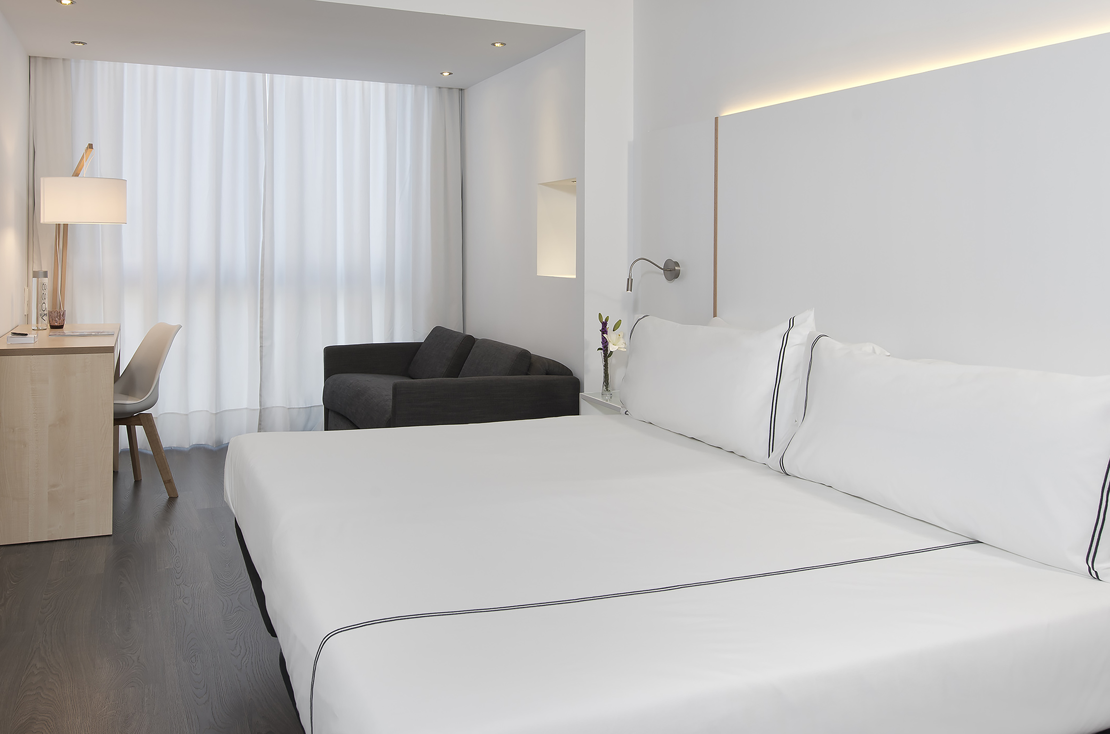 Modern hotel room with white bed, gray sofa, desk, and bright minimalist decor.