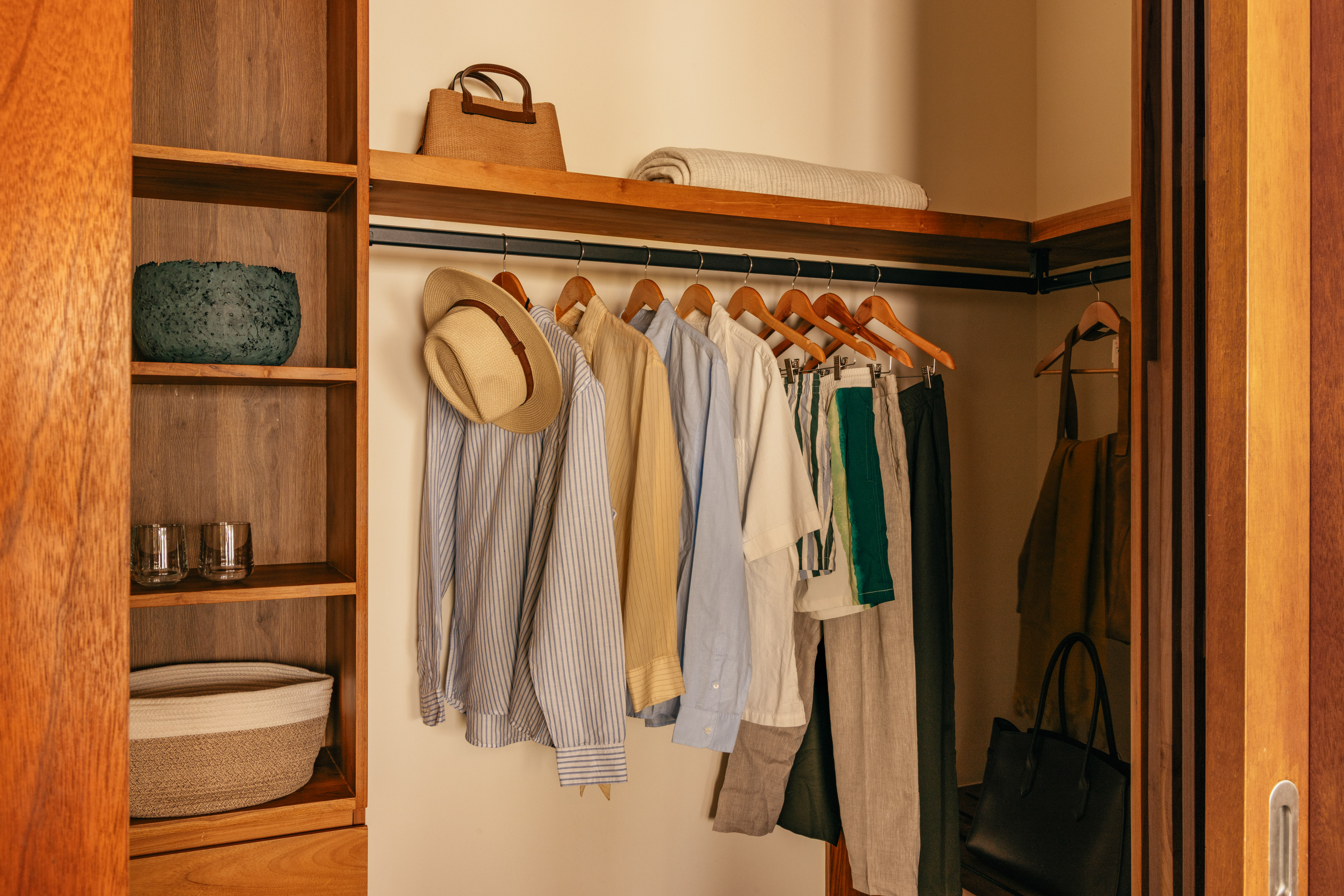Organized wooden walk-in closet with hanging clothes, hat, and storage.