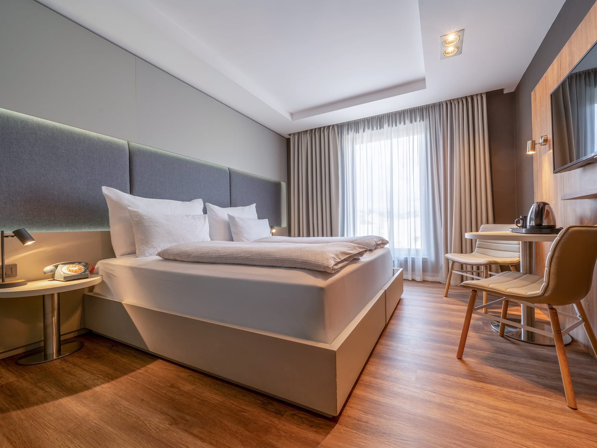 Modern hotel room with comfortable bed, wood floor, bright window, and seating area.