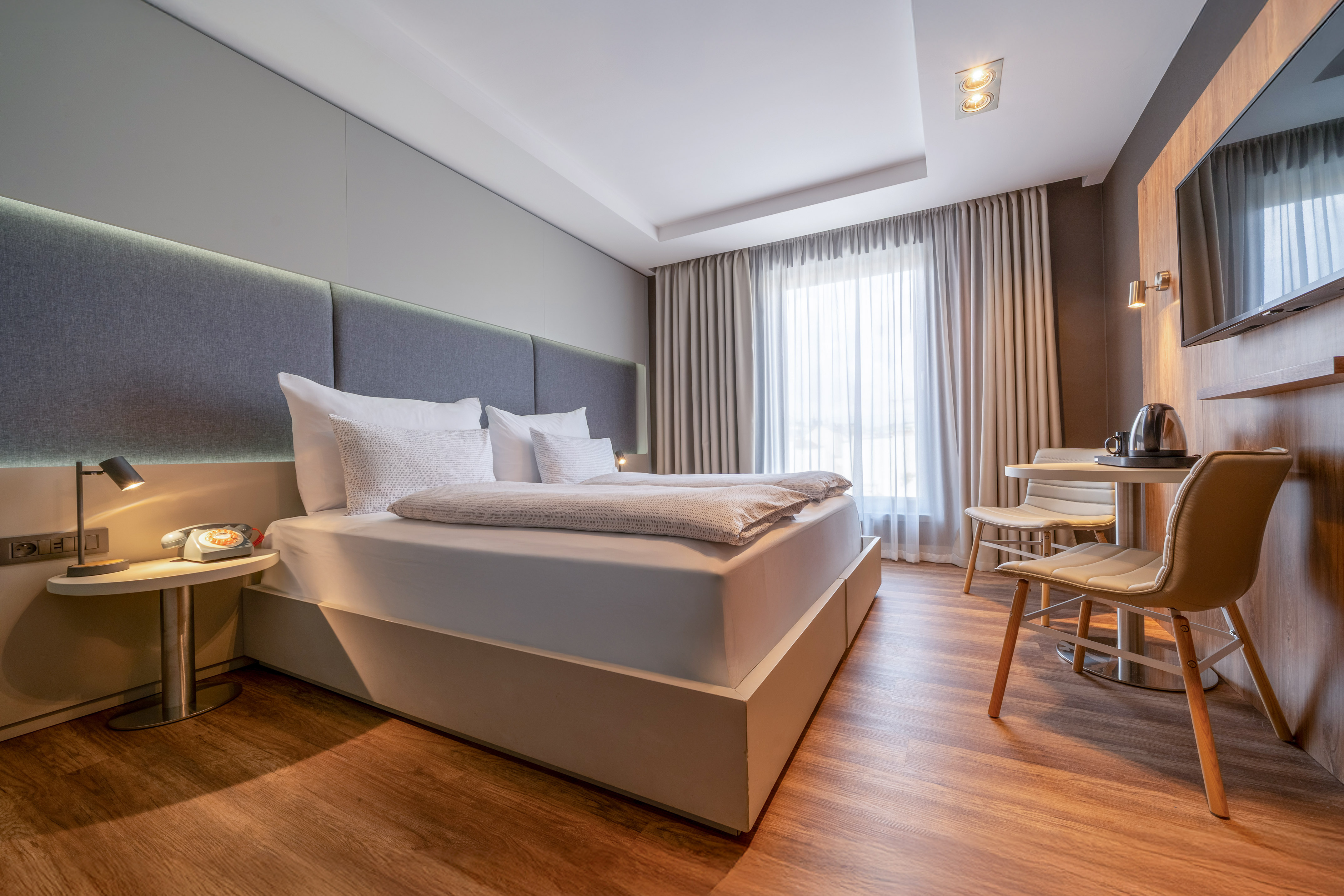 Modern hotel room with comfortable bed, wood floor, bright window, and seating area.