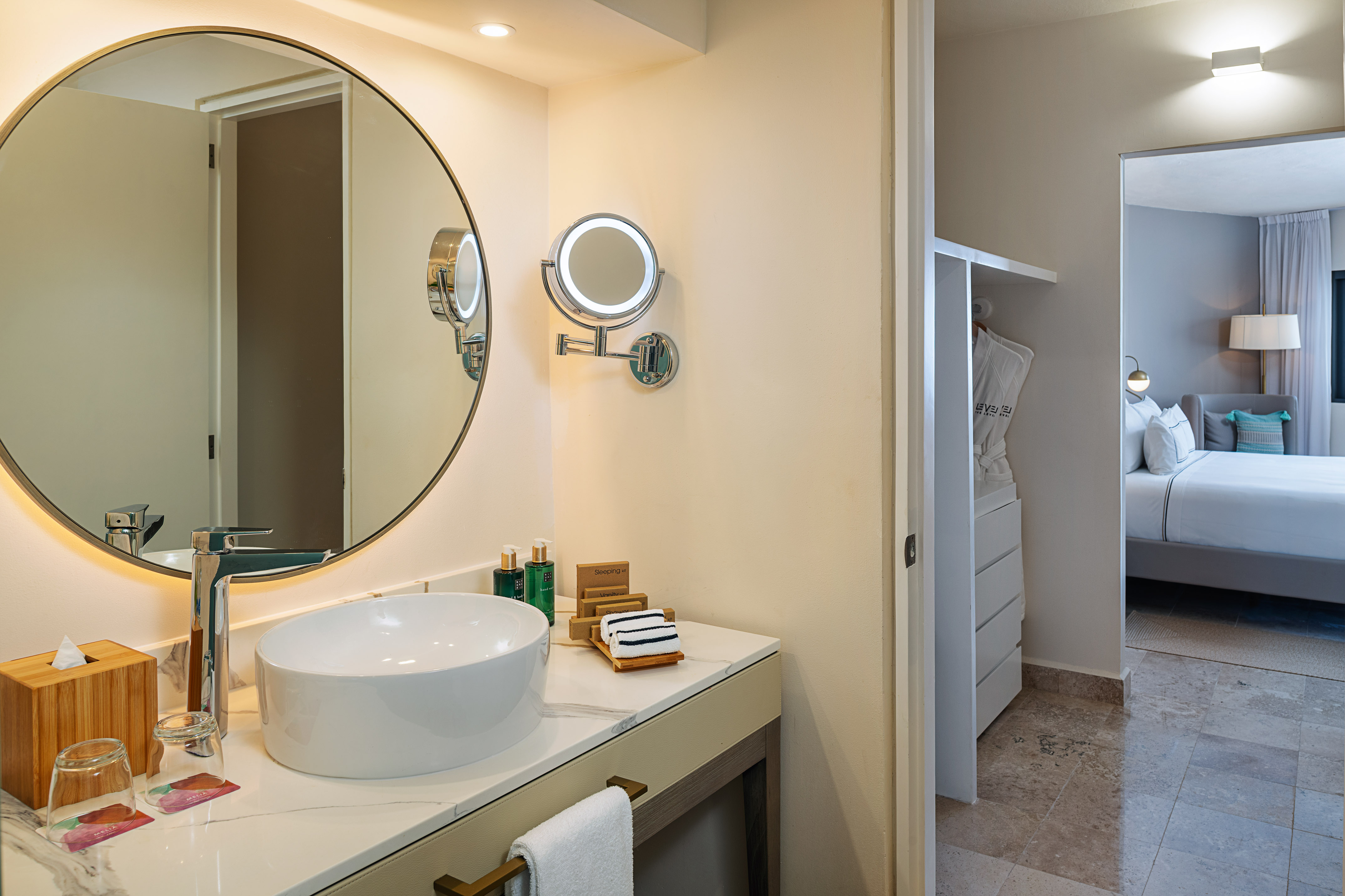 Modern hotel suite bathroom vanity with round mirror and connecting bedroom.