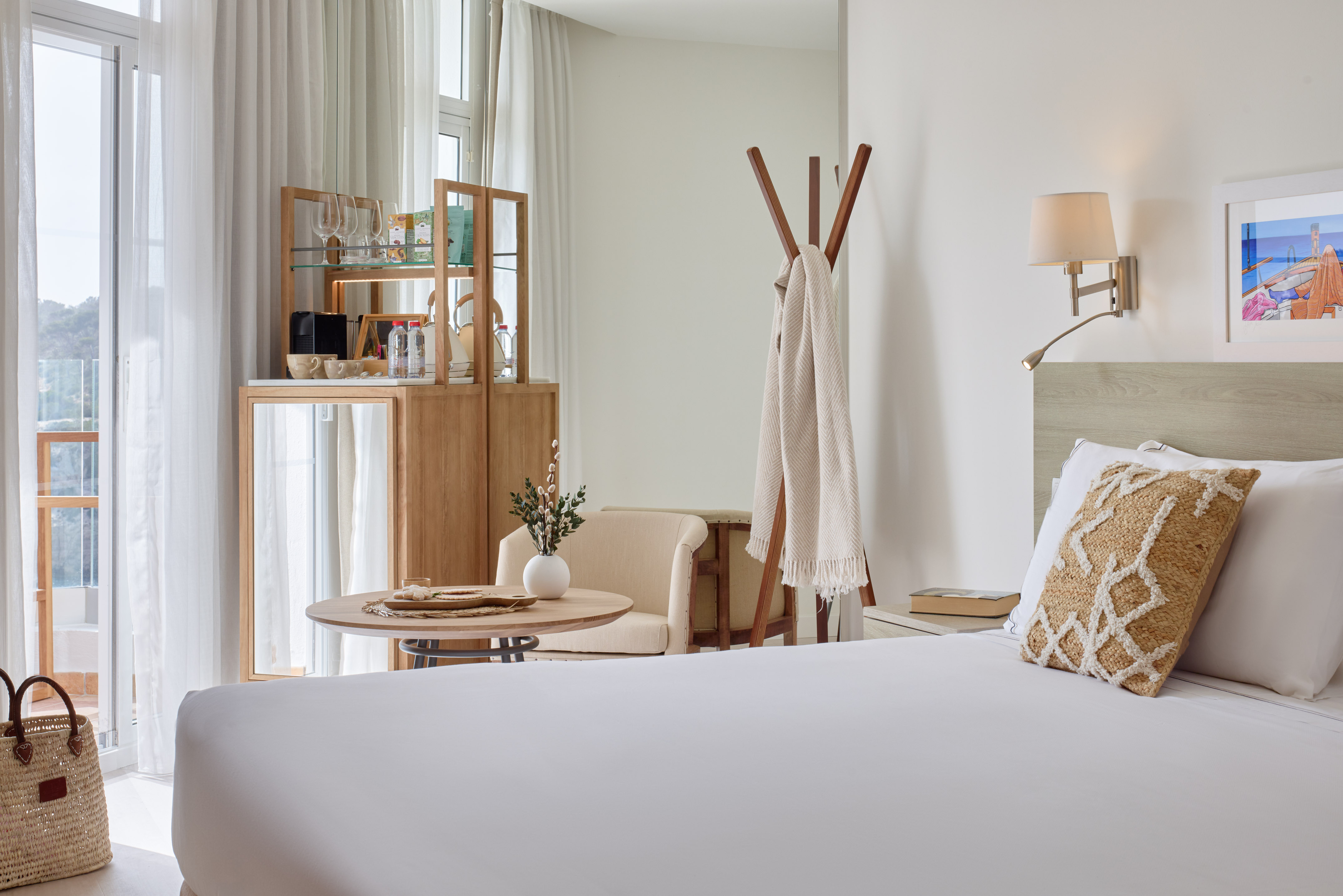 Bright modern hotel room with white bed, balcony view, mini-bar, and wooden decor.